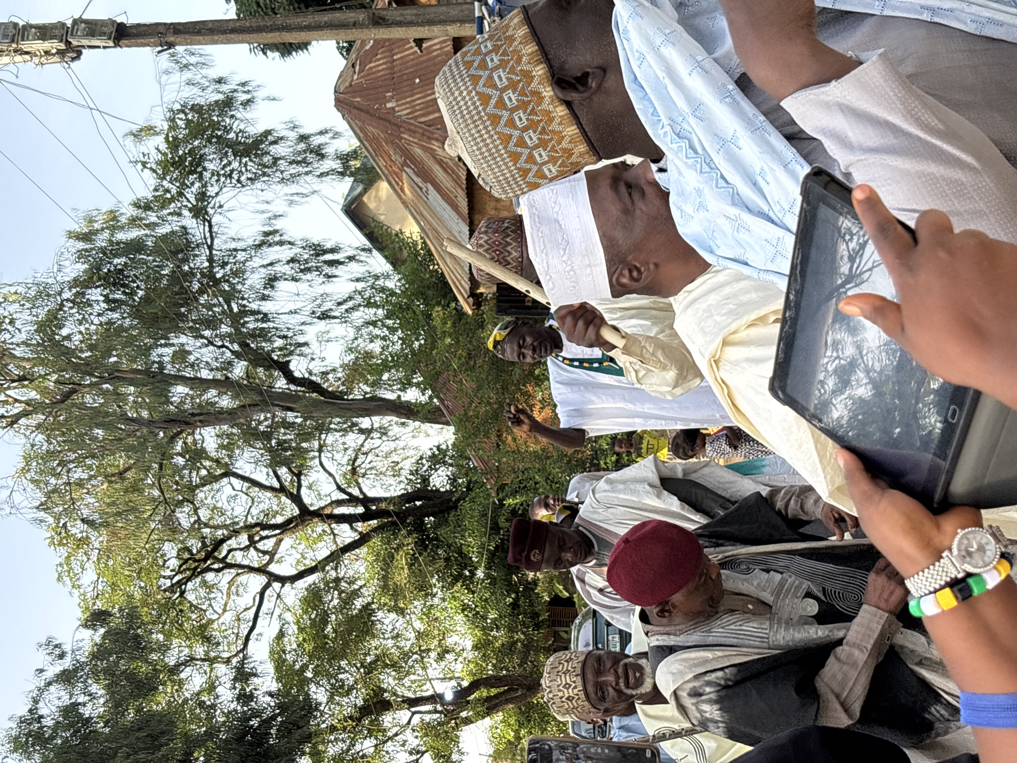The image depicts a gathering of individuals, primarily men, dressed in traditional attire. Many are wearing kufi caps of various designs and colors, including white, patterned brown and white, and solid red. Some are also adorned in flowing robes and kaftans. The scene appears to be outdoors, with lush green trees and foliage visible in the background, suggesting a possibly tropical or subtropical environment. A corrugated iron roof of a building is also visible, hinting at a semi-urban or rural setting.
In the foreground, a hand is holding a tablet device, capturing an image or video of the scene. The tablet screen shows a reflection of the trees. The hand is also wearing a wristwatch with a beaded bracelet featuring green, yellow, and black colors. Another hand, wearing a blue band, is also visible near the tablet.
The expressions on the faces of the men are varied, with some looking towards the camera or the person taking the photo, while others appear to be in conversation or observing something else. The atmosphere seems to be one of a community event or gathering, possibly a religious ceremony, celebration, or important meeting. The lighting suggests it is daytime, with clear skies.
While no explicit text is visible in the image, the attire and the gathering suggest a cultural event, potentially in Jos, Nigeria, given the provided location context. The overall impression is one of community and tradition.