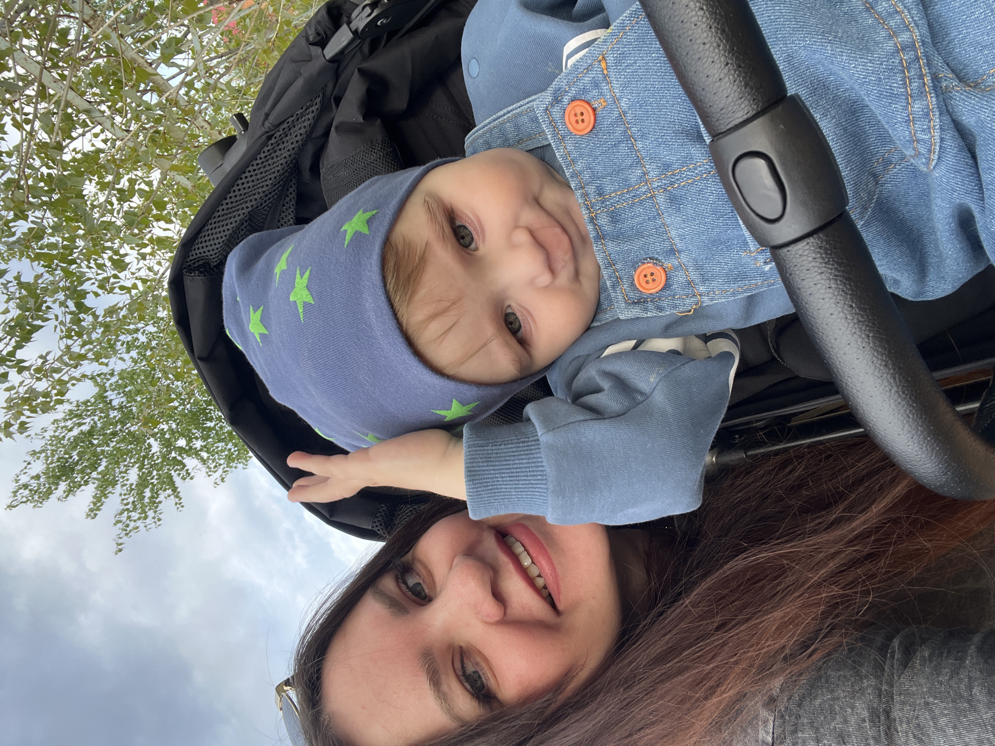 A photograph taken outdoors shows a woman and a baby. The woman is in the foreground with her head and upper body visible. She has long, brown hair, fair skin, and is smiling. The baby is in a stroller above the woman, wearing a blue hat with green stars and a blue denim jacket. The baby is also smiling. The stroller is black. In the background, there are trees and a cloudy sky. The location is not possible to determine from the given image.