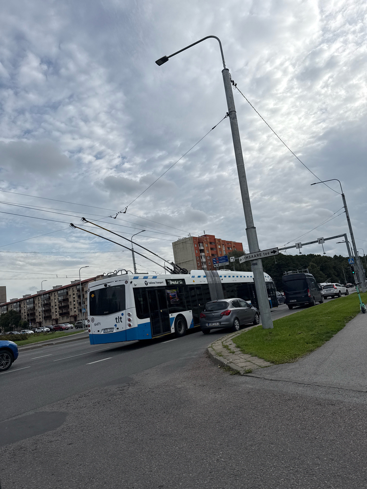 The image shows a trolleybus in Tallinn, Estonia, at an intersection with overhead wires. The trolleybus is white and blue, with "tlt" and "Tallinn Transport" branding. The license plate reads "015 TAK". A grey hatchback car is visible behind the trolleybus, with license plate "267 LVL". Other vehicles, including a black van and a white SUV, are further down the road. A street sign indicates "TAMMSAARE TEE" pointing to the right. Buildings line the left side of the road, and trees are visible in the background. The sky is cloudy.