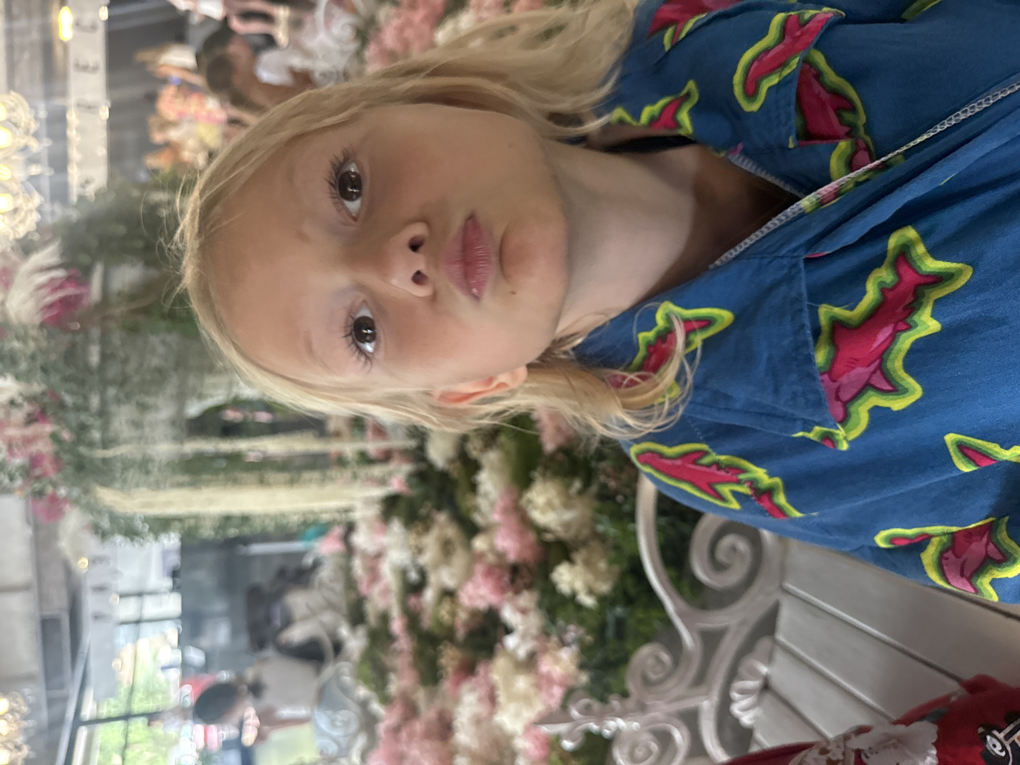 In the foreground of the image, a young girl with blonde hair is looking directly at the camera with a puckered expression. She is wearing a blue garment with a pattern of pink and green sharks. Behind her, a white ornate bench is visible, adorned with what appears to be large floral arrangements in shades of pink and white. The background is blurred but suggests an indoor setting with chandeliers hanging from the ceiling and other people in the distance. The overall impression is that of a festive or decorative event. The location is stated to be the city of Yalta, Ukraine.