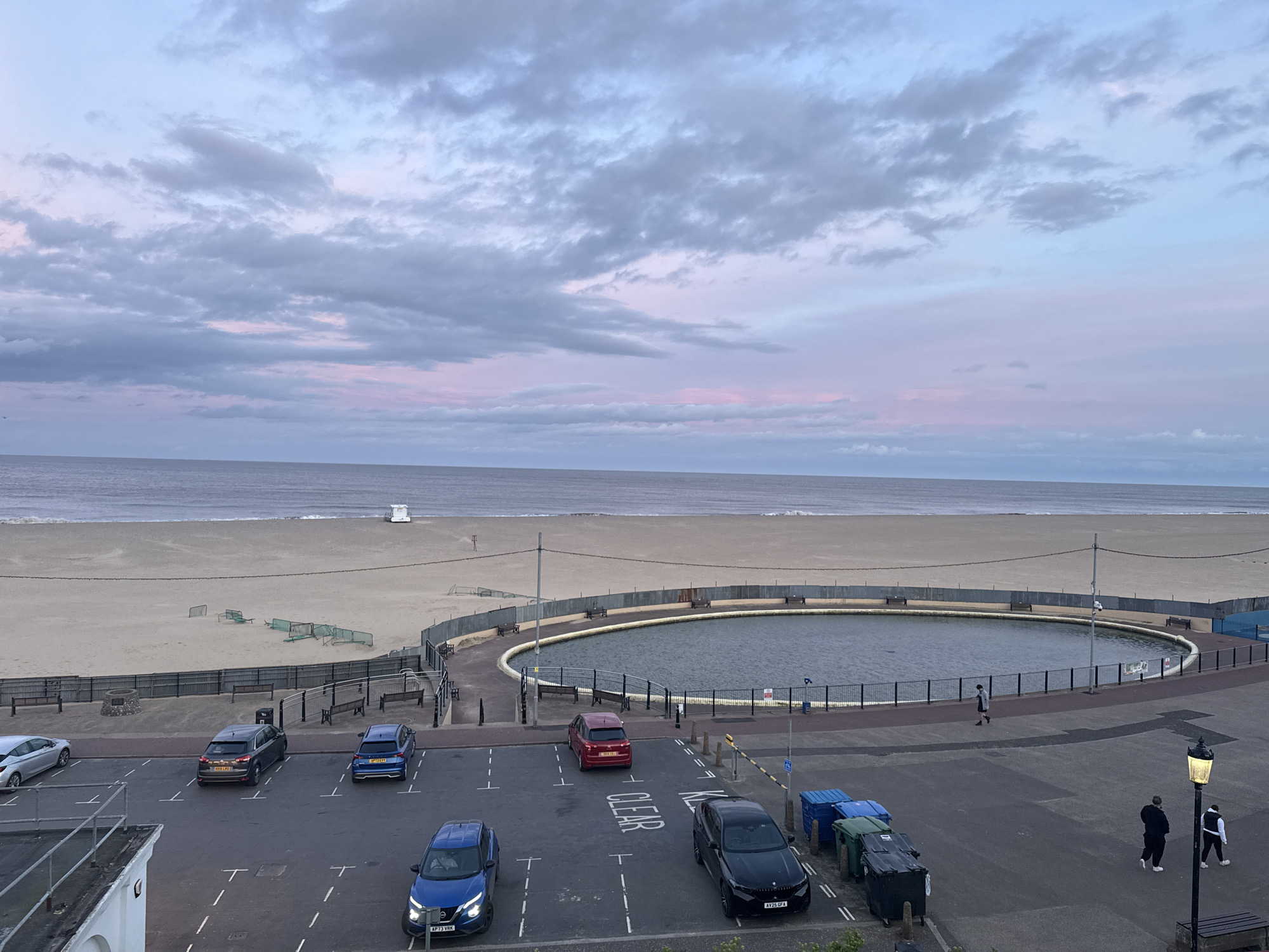 The image depicts a coastal scene in Gorleston-on-Sea, United Kingdom. A sandy beach stretches towards the horizon, with a lifeguard tower visible in the distance. A circular paddling pool, enclosed by a fence, is positioned prominently near the seafront. A parking lot separates the pool from the building the photo was taken from, containing several parked cars: a silver hatchback, a dark grey crossover, a blue hatchback, a red hatchback, and a black sedan, along with several trash and recycling containers. The parking lot is marked with white lines. Two people are walking on the pavement to the right, near a vintage street lamp. The sky is overcast with pink tinged clouds.