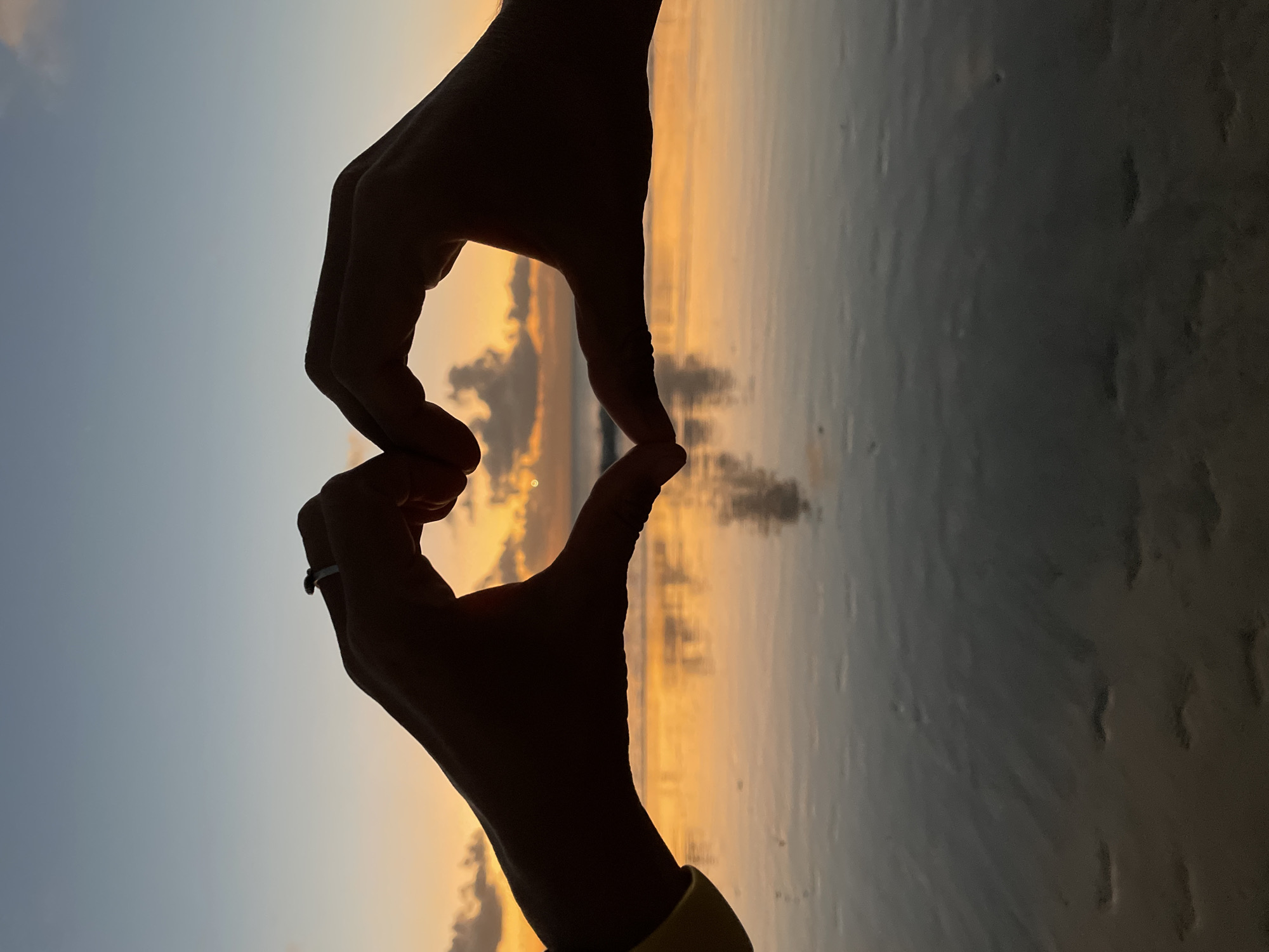 At sunset, two silhouetted hands form a heart shape against a colorful sky over the ocean in Kuta Selatan, Indonesia. The sky transitions from a soft blue at the top to vibrant oranges and yellows near the horizon, with a few scattered clouds. The ocean reflects the warm hues of the sunset, creating a shimmering surface. The foreground shows a dark, sandy beach with the wet sand reflecting the light. One of the hands is wearing a ring.
