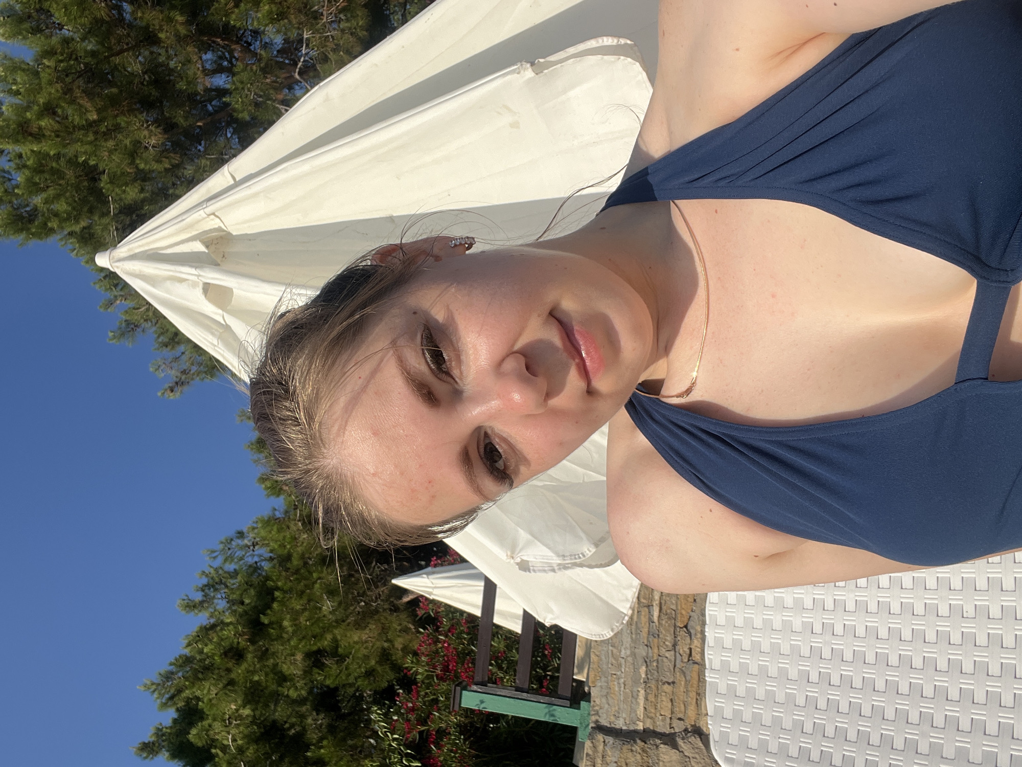 A young woman with blonde hair pulled back is taking a selfie outdoors. She is wearing a dark blue halter-neck swimsuit and a thin gold necklace. Her skin has a slightly tanned appearance with some redness, possibly from sun exposure. She is smiling slightly and looking towards the camera.
Behind her, a large white beach umbrella is partially visible, its fabric creased and casting shadows. The background includes lush green trees and a clear blue sky. To the left of the woman, there is a section of a stone wall and a wooden fence with green posts, adorned with red flowers. The woman is seated on a white lounge chair with a distinctive criss-cross pattern. The scene suggests a sunny day at a beach resort or similar outdoor recreational area in Alanya, Turkey.