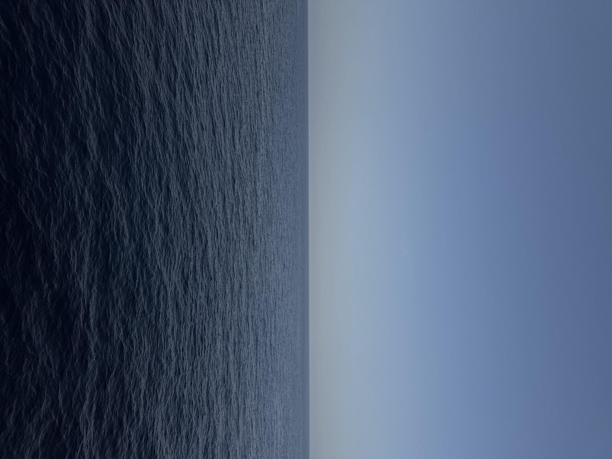 The image depicts a vast expanse of dark blue ocean meeting a clear, pale blue sky at a sharp, unbroken horizon line. The water exhibits a texture of small, consistent ripples, suggesting a calm but dynamic surface. The sky above is featureless, with a subtle gradient from a lighter shade near the horizon to a slightly deeper blue further up. No landmasses, vessels, or living creatures are visible. The overall impression is one of immense space and stillness. The location is given as Unknown, Unknown.