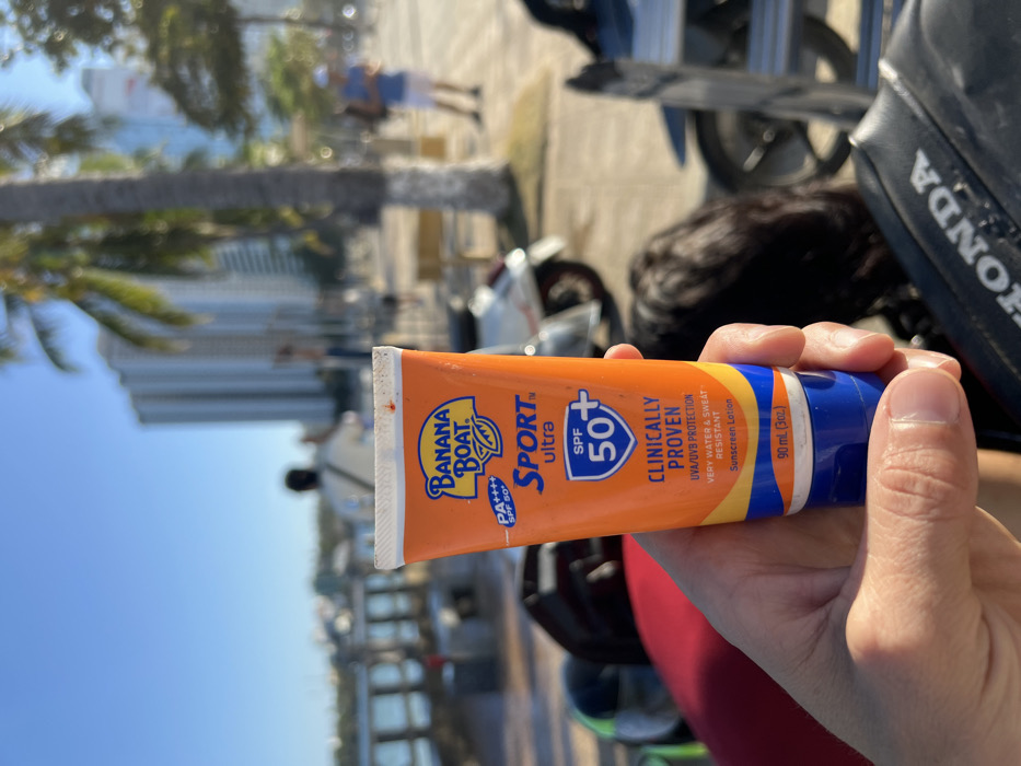 A light-skinned hand holds an orange, blue, and yellow tube of Banana Boat Sport Ultra sunscreen, SPF 50+, PA++++, 90 ml (3 oz), in the foreground. The tube's label prominently displays text including "CLINICALLY PROVEN," "UVA/UVB PROTECTION," and "VERY WATER & SWEAT RESISTANT." The fingers show visible knuckles and trimmed fingernails.
The background, rendered with a shallow depth of field, depicts a sunny outdoor environment on a paved promenade in Nha Trang, Vietnam. To the immediate left of the hand, a fragment of red fabric and a dark helmet are visible. Below the hand, a black motorcycle seat bearing the "HONDA" logo is partially present. Further back, several palm trees line the promenade. Multiple individuals are present: one person, viewed from behind in a light-colored top, stands with an arm raised to the left; another person in a blue shirt and white shorts stands to the right, appearing to interact with a handheld device. A light-colored scooter is parked on the walkway. In the far left background, a body of water with a pier or embankment structure is visible. The distant horizon features numerous tall, modern high-rise buildings under a clear blue sky, indicative of the Nha Trang coastline.