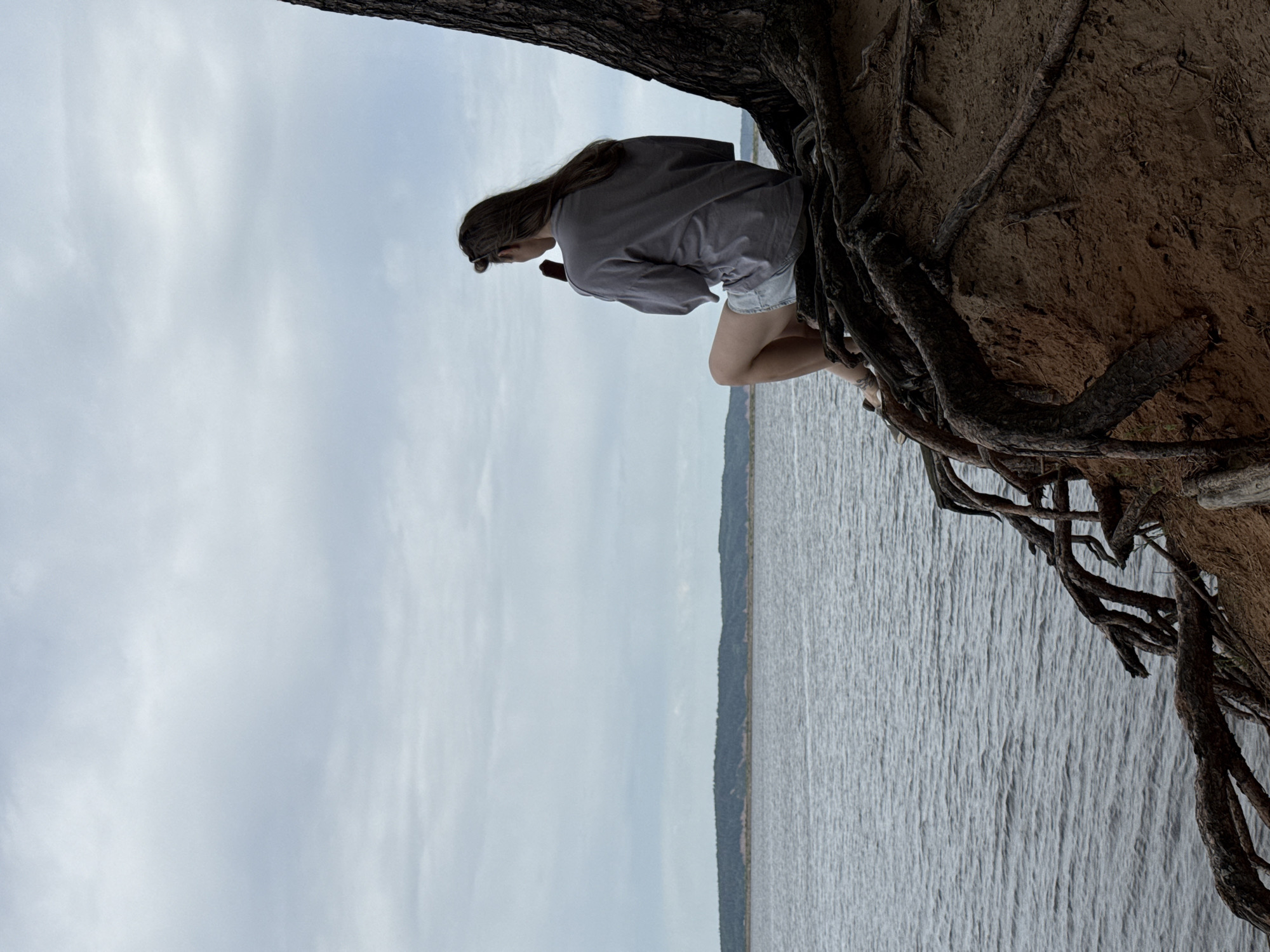 A lone woman with long brown hair, wearing a grey long-sleeve shirt and denim shorts, sits precariously on the exposed roots of a large tree overlooking a wide body of water. The water reflects a grey, cloudy sky. The roots of the tree are tangled and exposed, growing out of a sandy embankment. In the background, a distant treeline can be seen on the opposite shore of the body of water. The scene appears to be taken at a park or natural area near the city of Chaikovsky, Russia.