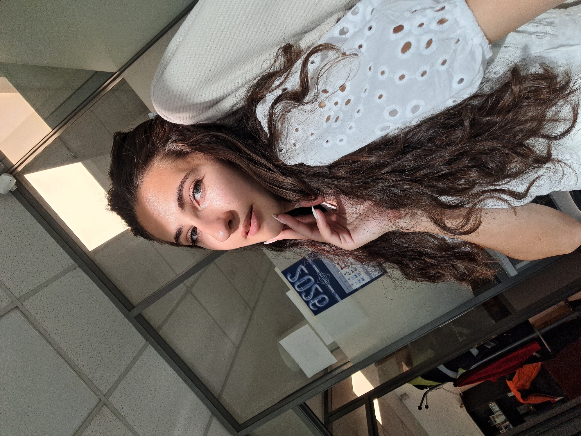 The image shows a young woman with long, wavy brown hair, wearing a white eyelet-style top. She is posing for a selfie, with her left hand resting on her chin and her right arm slightly visible. She appears to be indoors, possibly in an office environment, with a glass partition and a ceiling with acoustic tiles and fluorescent lighting visible in the background. A calendar with the ASOS logo is also visible on the glass partition. The scene is lit by a combination of overhead lighting and natural light, casting shadows on the wall and ceiling.