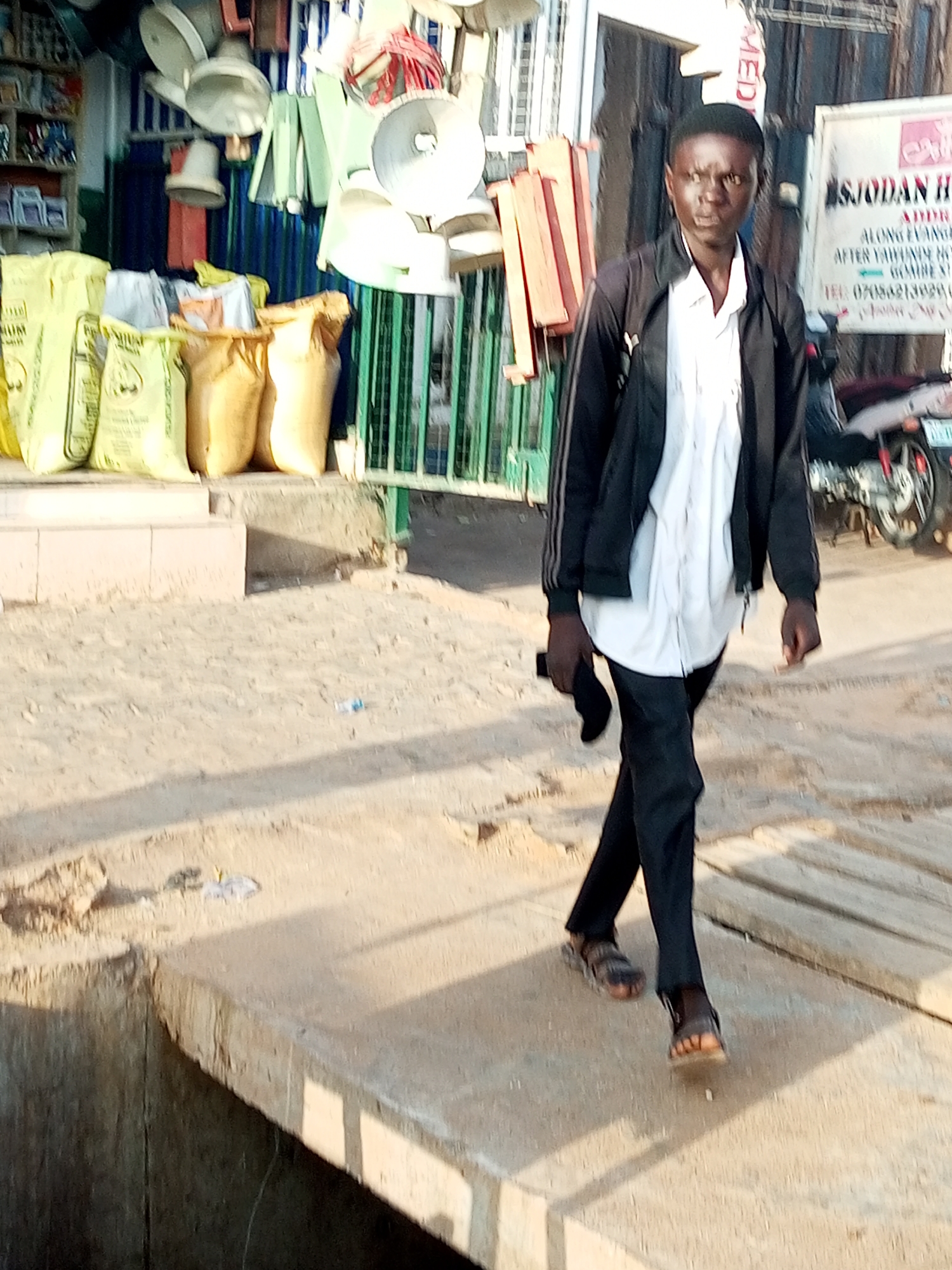 A young man walks along a sandy pathway in what appears to be a market area. He is wearing a black jacket with white stripes on the sleeves over a white shirt and black trousers. He has on sandals and is carrying a dark-colored object, possibly a phone, in his left hand. The man's gaze is directed forward, and his expression is neutral.
In the background, there are stalls displaying various items, including what look like large sacks, possibly agricultural produce, stacked in yellow and tan bags. There are also what appear to be electrical appliances like fans and light fixtures hanging from above. A green metal fence runs along the stalls. A motorcycle is parked to the right of the man.
A sign is visible on a building in the background with text that reads "SJODAN HO" at the top, followed by "ALONG GAMBERI ST.", and "AFTER YAHUADE NIGERIA". Below that, there is a phone number: "TEL: 07068213928". Another line of text below the phone number is partially visible.
The lighting suggests it is daytime, possibly late afternoon, given the long shadows cast on the ground. The weather appears to be dry. The scene is consistent with a busy market setting in Gombe, Nigeria.