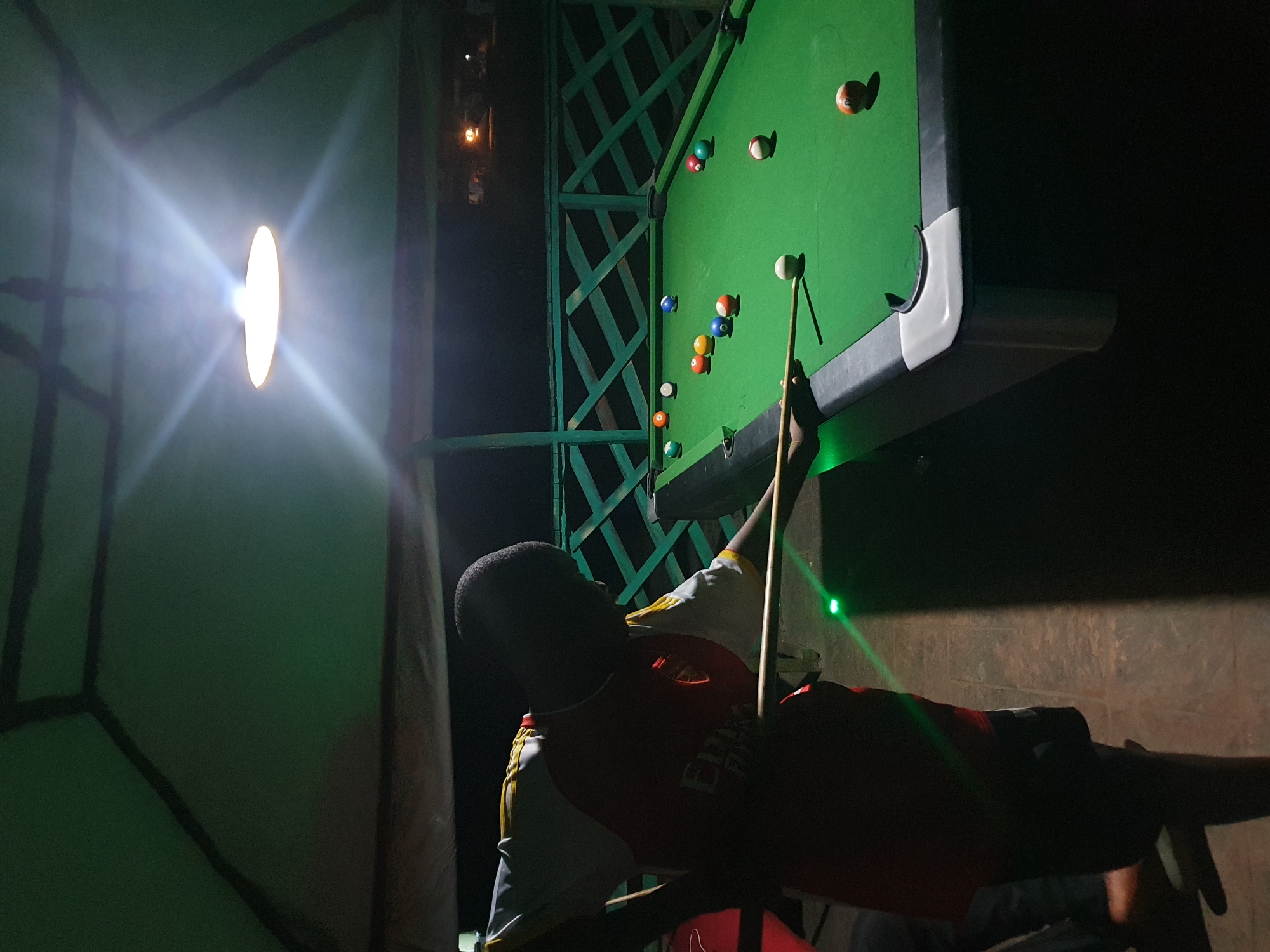 In Lokoja, Nigeria, a person is playing pool at night. The scene is set under a green, tent-like structure with a bright light overhead. The pool table is green, with several balls scattered across it. The player is wearing a red and white shirt and is in the process of hitting a white ball with a cue stick. A green laser light is visible on the floor. The background shows a wooden fence and the night sky, with a distant light source.