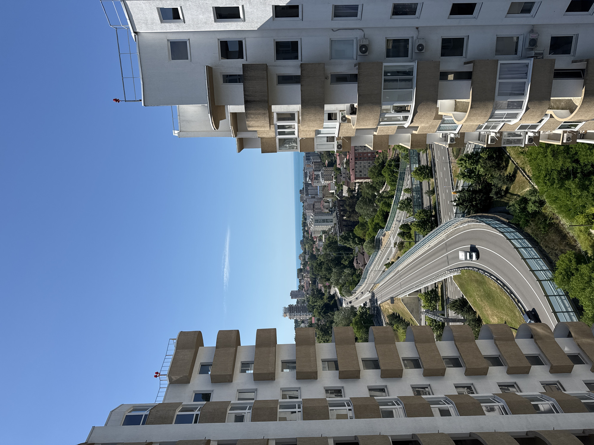 The image is an aerial view of a city with a highway curving through it, leading towards the ocean. Two tall, modern apartment buildings with numerous balconies are prominently featured on the left and right sides of the frame. The buildings are white with beige balconies. The highway is a multi-lane road with a median and sound barriers, and there are a few vehicles visible on it. Beyond the highway, a dense urban landscape with more buildings of varying heights and architectural styles can be seen, interspersed with lush green trees. In the distance, the calm blue sea stretches to the horizon under a clear, bright blue sky. This scene is characteristic of the Khosta district in Sochi, Russia, a popular resort city on the Black Sea coast. The image captures a sunny day, with strong shadows indicating the time is likely midday. There are no people directly visible in the scene.