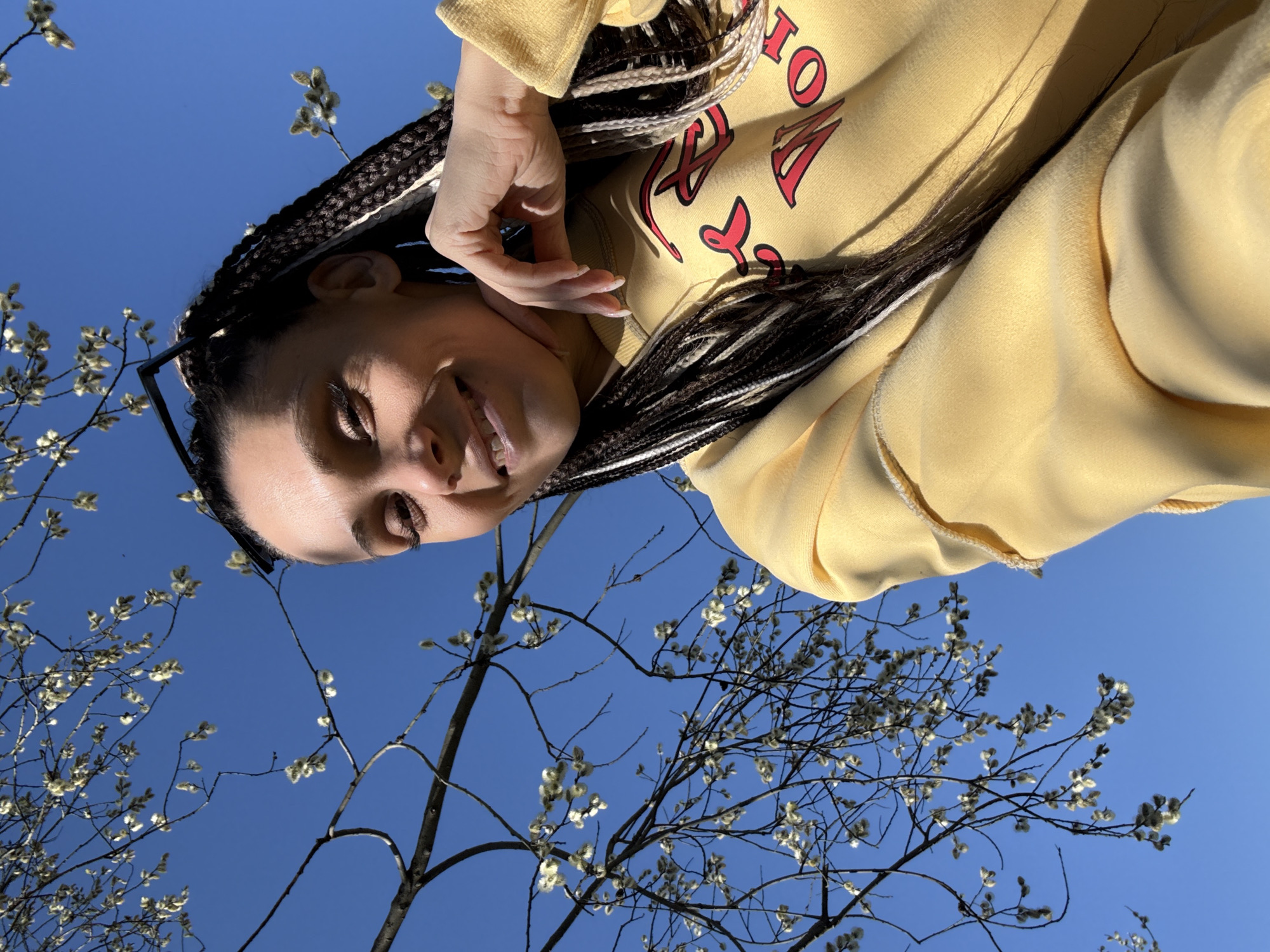 The image shows a person in Unknown, Unknown, wearing a yellow sweater, and looking upwards. The sweater has red letters on it. The person has braids with black and white strands. They are holding their hand near their face. The background features a clear blue sky and budding tree branches.