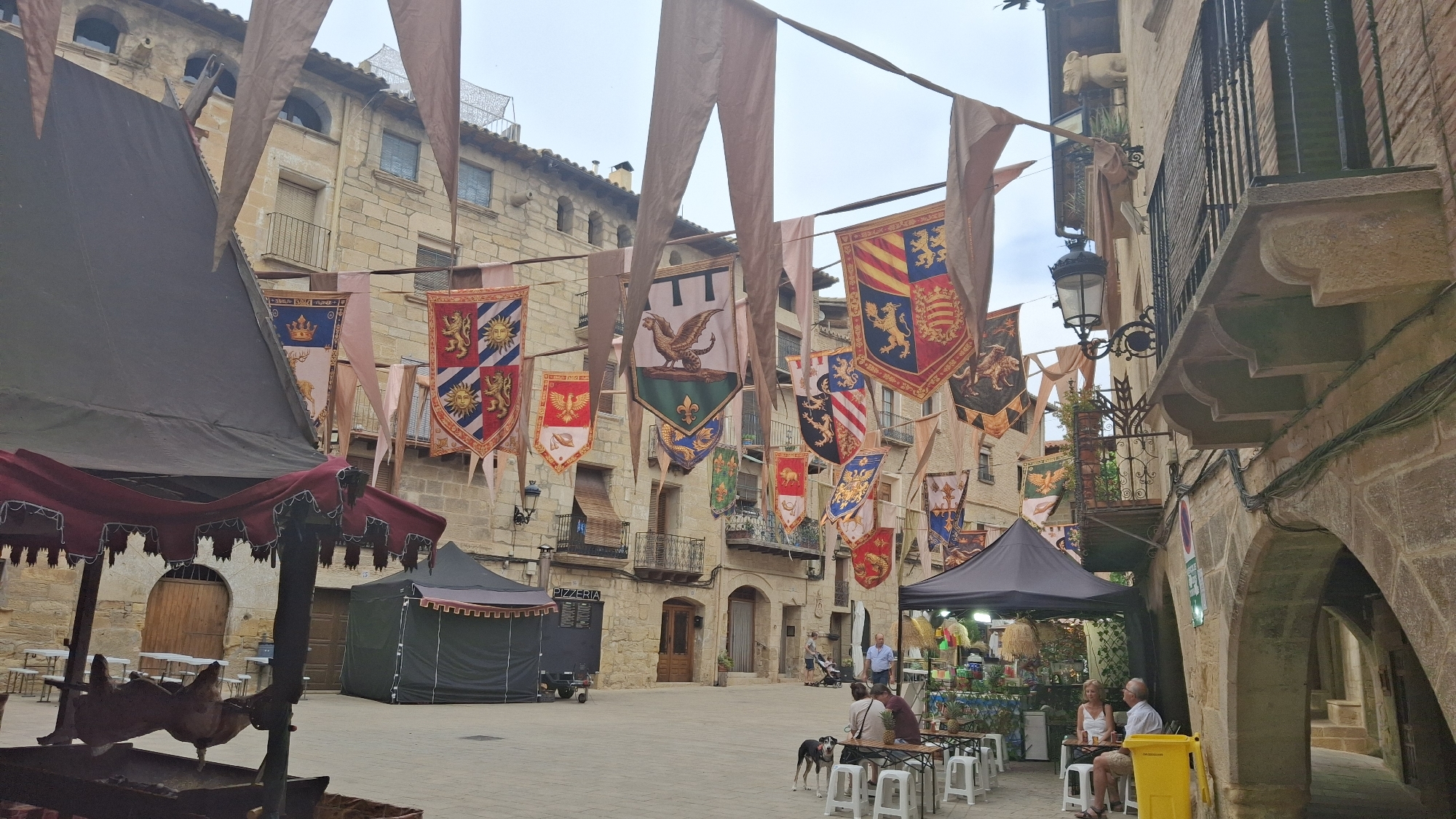 The image depicts a street scene in La Fresneda / la Freixneda, Spain, likely during a medieval-themed festival. The foreground features a large, dark-colored tent with a burgundy trim and decorative fringe, under which are two tables with white stools. A spit roast with what appears to be poultry is visible in the lower left corner. Beyond this, a paved square opens up, with other tents and market stalls set up. Colorful medieval banners, depicting various crests and heraldic symbols such as lions, eagles, and suns, are strung across the street, adding to the festive atmosphere.

The background is dominated by traditional stone buildings with arched windows and balconies. Some of the buildings have a worn, aged appearance, characteristic of historical architecture. People are scattered throughout the scene. In the mid-ground, a few individuals are seated at tables outside a tent labeled "PIZZERIA". Further back, two men and a woman are seated at a table, with a dog nearby. Other people are seen walking in the distance. The sky is overcast, suggesting a cloudy day. The overall impression is one of a bustling, historical town square transformed for a special event.