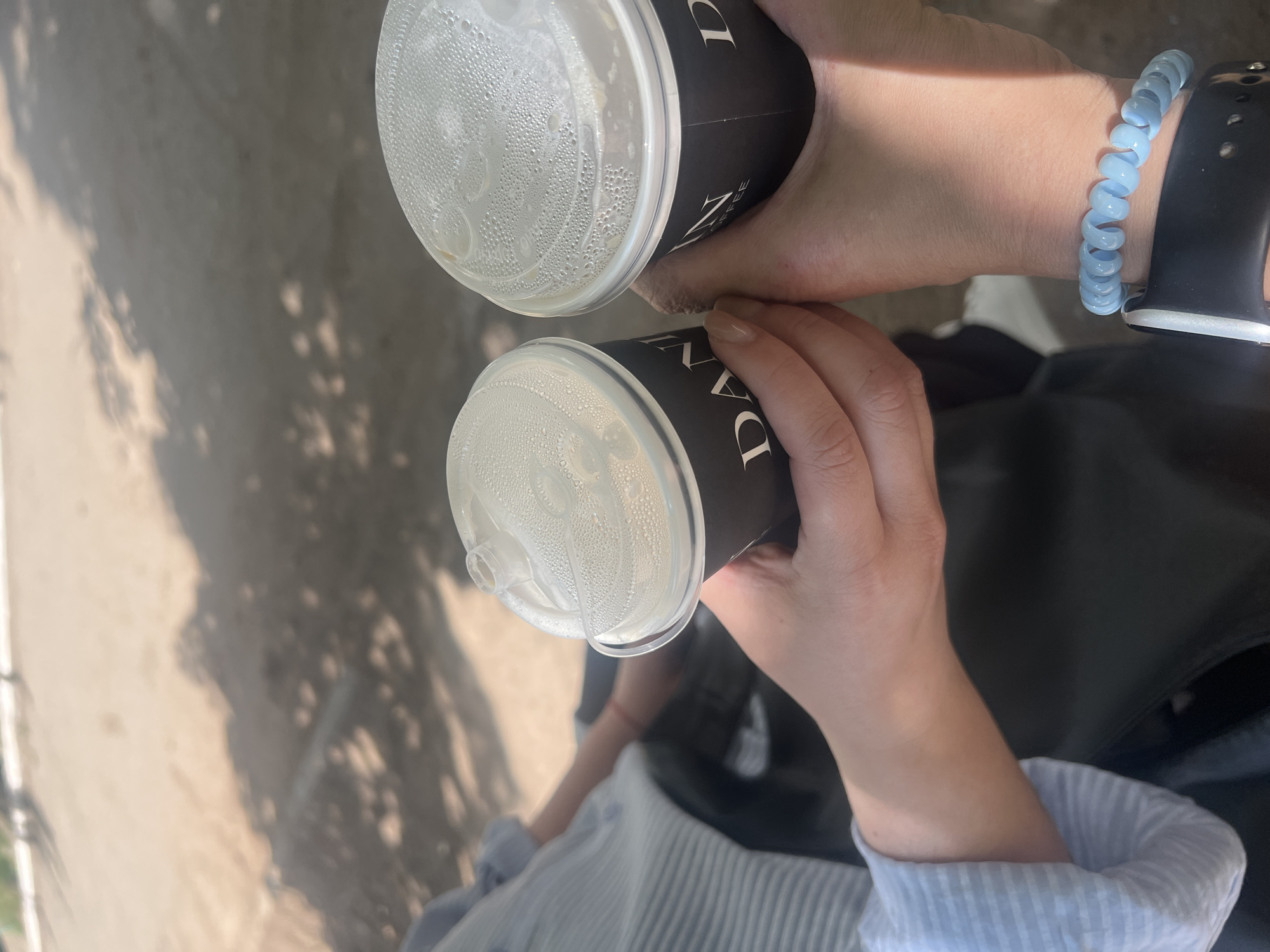 Two people are holding coffee cups. The cups are black with white lettering that says "DAN COFFEE". The cups have clear plastic lids covered in condensation. The background is a sandy area with shadows cast from trees. One person is wearing a light blue striped shirt, and the other is wearing a black jacket. The person on the right is wearing a light blue spiral bracelet on their wrist and a black smartwatch. The image is taken outdoors during the day, likely in an urban park or outdoor seating area of a cafe.