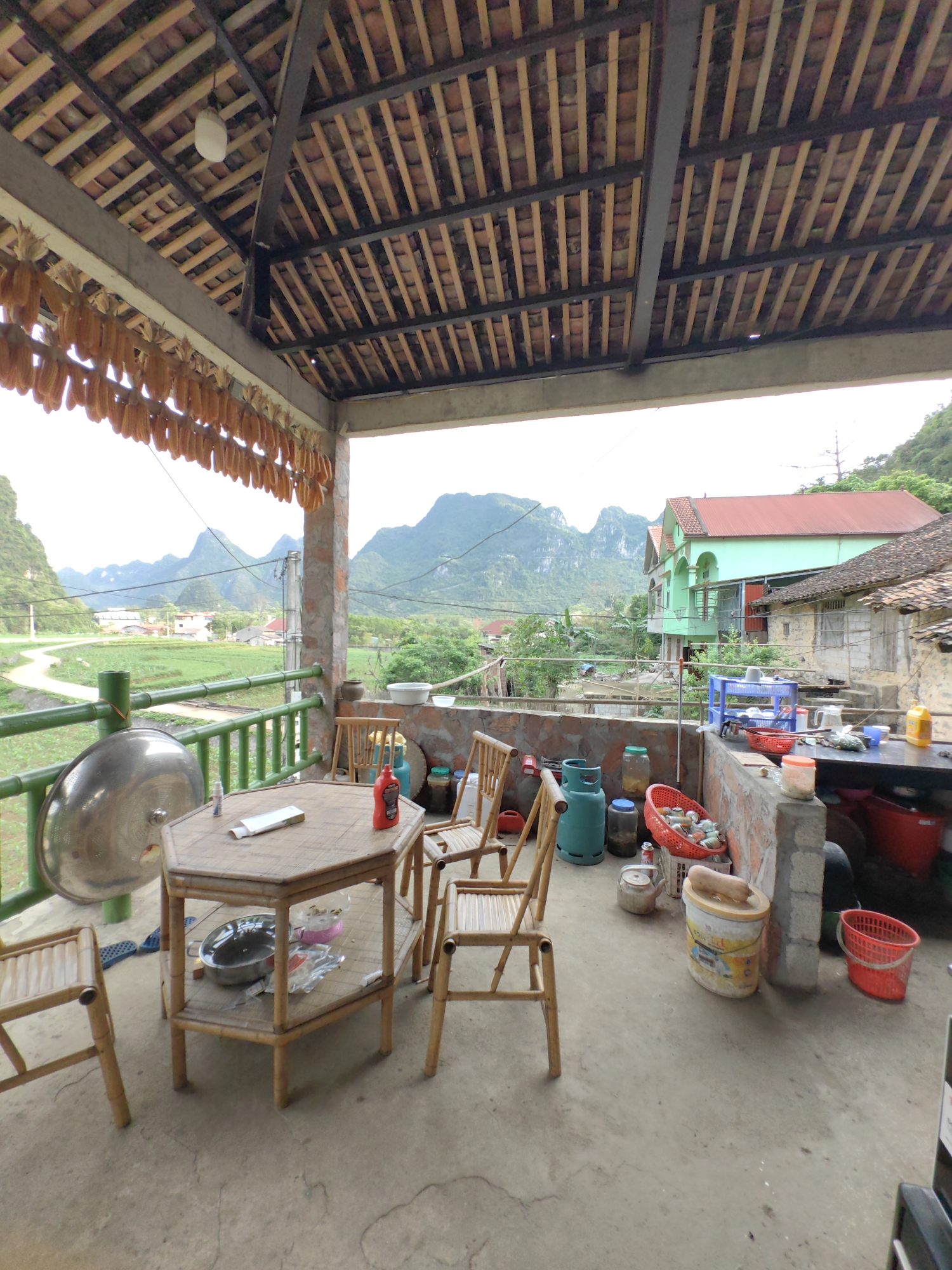 The image shows a covered patio in Xã Đàm Thủy, Vietnam, with a rustic aesthetic. Corn hangs along the edge of the roof. A hexagonal table with bamboo chairs sits on the patio, along with a variety of household items, including propane tanks, pots, buckets, and cooking utensils. In the background, there is a view of a rural landscape with mountains, fields, and houses. One house is green with a red roof, while others appear more traditional. A road or path is visible in the distance.