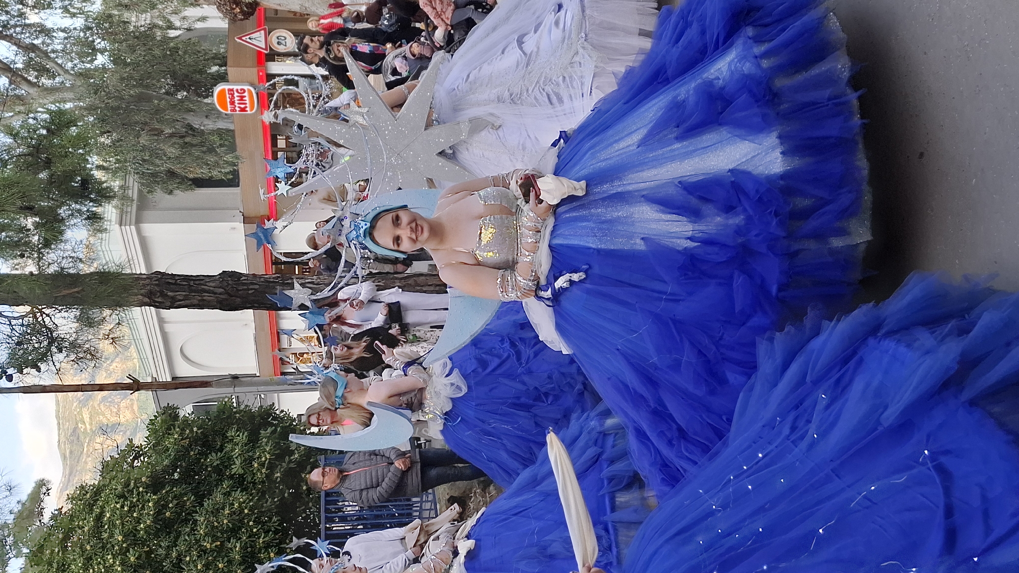 The image captures a vibrant scene of a carnival procession, likely in Budva, Montenegro. The focal point is a woman in an elaborate costume: a blue tulle dress with white accents, a silvery, star-shaped headdress adorned with blue stars, and a moon-shaped structure behind her. She is smiling and appears to be interacting with the crowd.
Behind her, another woman is similarly dressed in a white tulle dress with an identical headdress. In the background, a diverse crowd of onlookers is visible, observing the procession. There are trees and buildings that are likely located in Budva, Montenegro. A Burger King sign is visible in the background.
