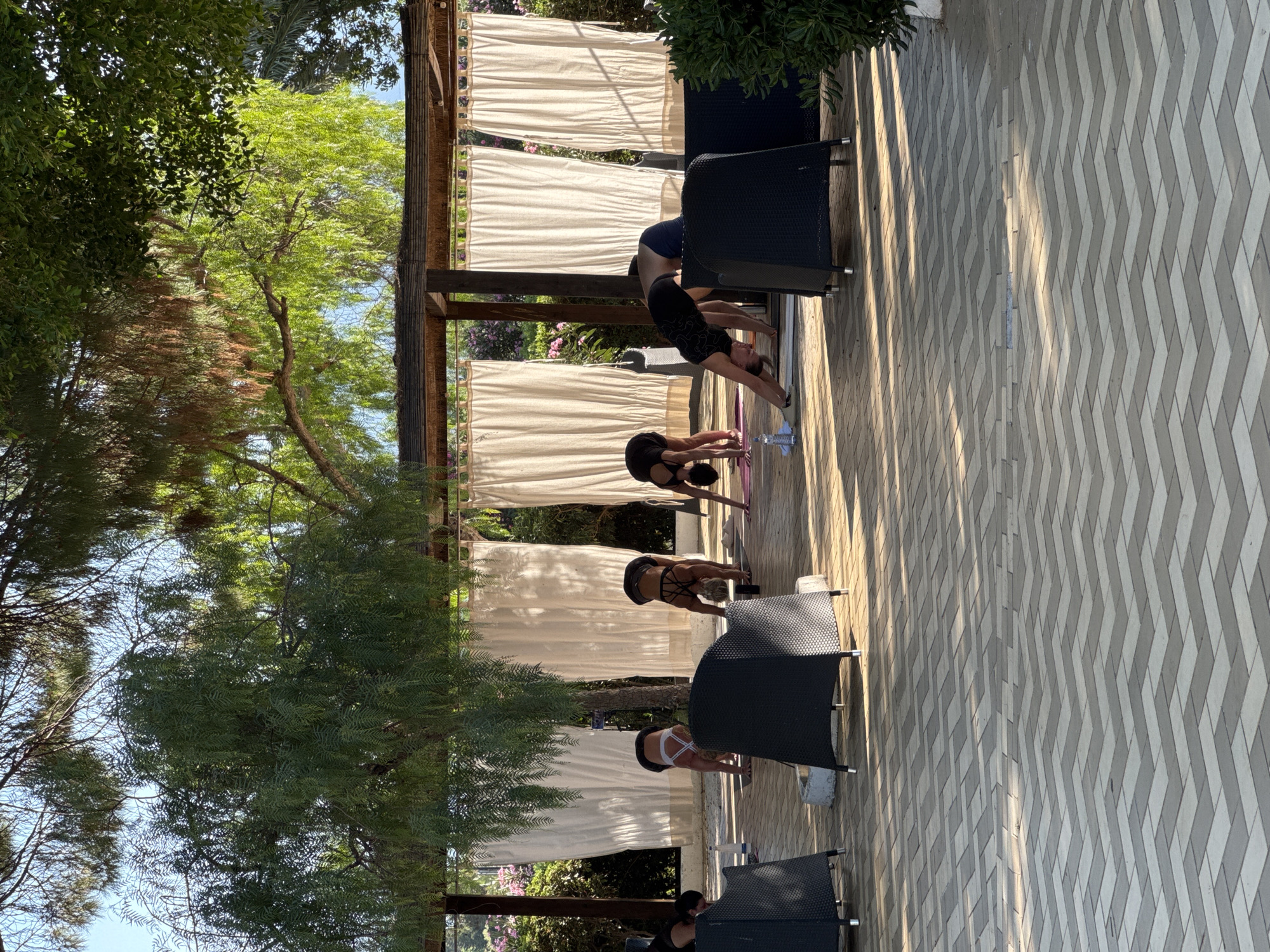 The image depicts an outdoor yoga class taking place under a wooden pergola covered with thatch. Several individuals, dressed in athletic wear, are engaged in various yoga poses on mats spread across a patterned paved patio. Behind the participants, white curtains hang, obscuring the background. The scene is set amidst lush greenery, with trees providing shade over the area. The overall ambiance suggests a serene and tranquil environment, possibly part of a resort or wellness center in Isola delle Femmine, Italy, given the architectural style and the Mediterranean vegetation. The patio flooring is composed of light and dark grey rectangular pavers arranged in a chevron pattern. Two black woven chairs are visible in the foreground.
