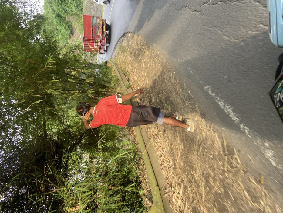 The image shows a man walking on a paved road in the city of Cipatat, Indonesia, at coordinates -6.799 latitude and 107.444 longitude. The man is wearing a red shirt and black shorts and is walking away from the camera. He is walking on the right side of the road, which is lined with a concrete curb. There is a small, dry ditch on the side of the road. The road is covered with dry leaves and other debris. There is a red truck in the background driving away from the camera and a silver car in the very far distance. The image appears to be taken from the back of a motorcycle. There is a green area with foliage on the left side of the frame. The man is not interacting with anyone else in the image. The sky is overcast.