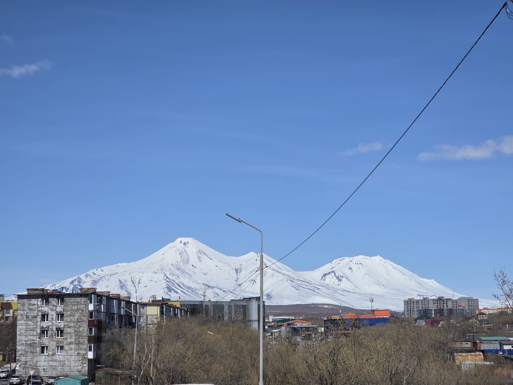 The image shows a city landscape with snow-covered mountains in the background, under a clear blue sky with sparse clouds. The foreground includes buildings of various heights and colors, some appearing older and others newer. Bare trees are visible, suggesting a transition between seasons. An electrical pole with wires stretches across the frame, slightly obscuring the view of the mountains. The overall setting appears to be a populated area in proximity to a mountain range. Location: Unknown, Unknown.