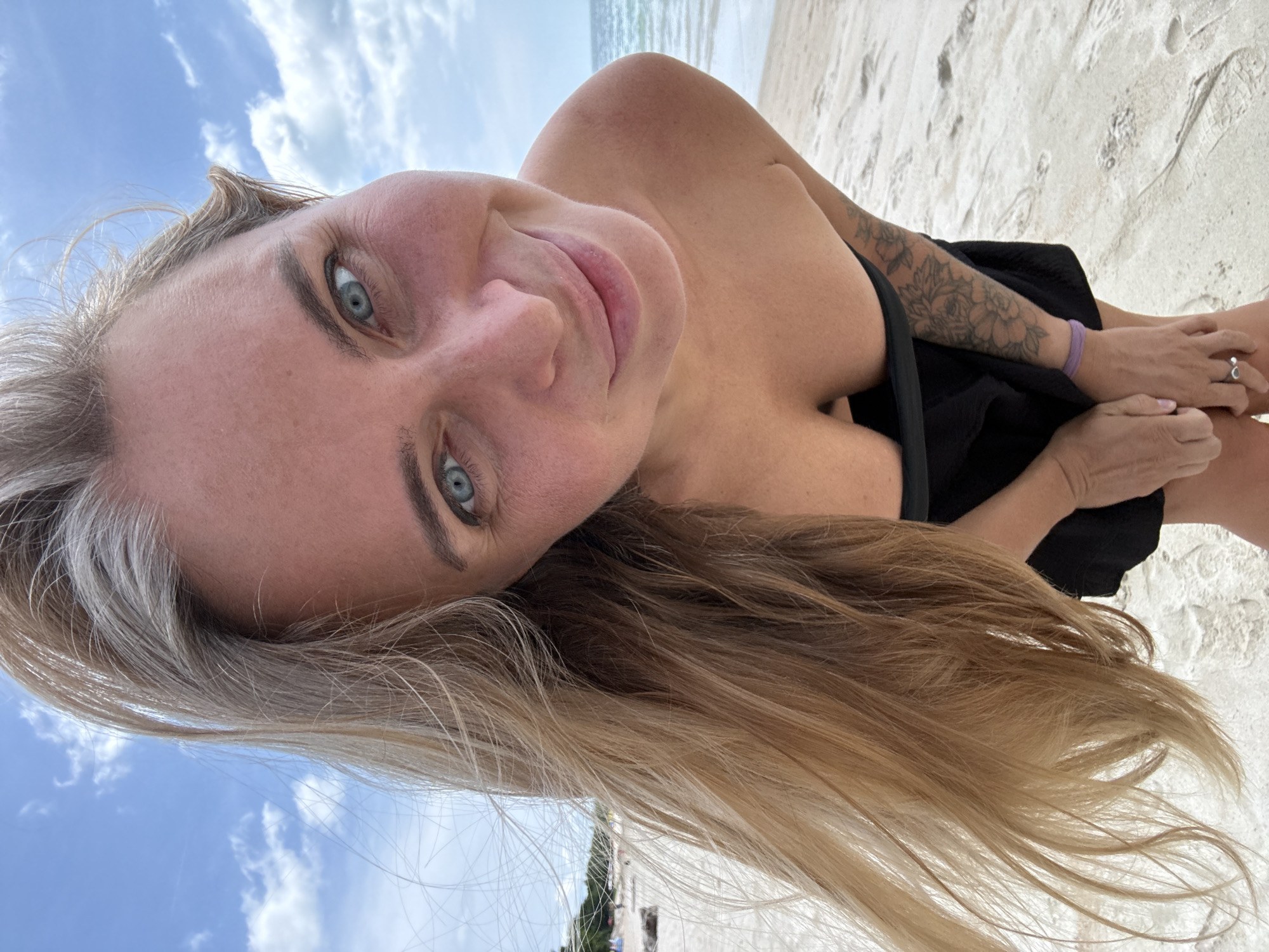 A woman with blonde hair and blue eyes is posing for a close-up photo on a beach. She is wearing a black top and has a tattoo on her arm. The background shows the ocean and the sky. The image appears to be taken in the Ko Samui District of Thailand.