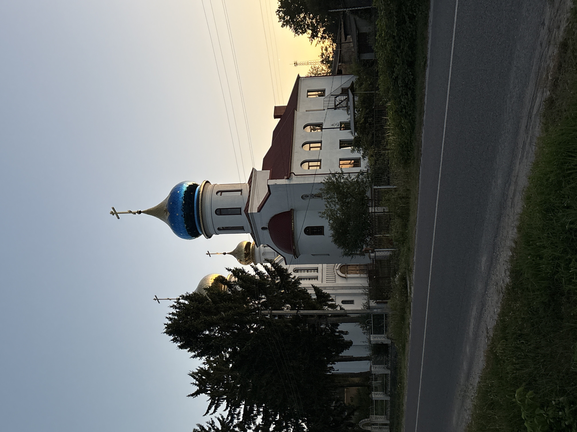 An Orthodox church with a blue and gold dome and multiple gold domes stands on the right side of the image, next to a road in Lanivtsi, Ukraine. The church is white with red trim and has several windows. A fence and trees are in front of the church, and the sky is a clear blue. The sun is setting in the background, casting a warm glow over the scene. The road in the foreground is paved and has a white line down the center. The area around the church is green and grassy.