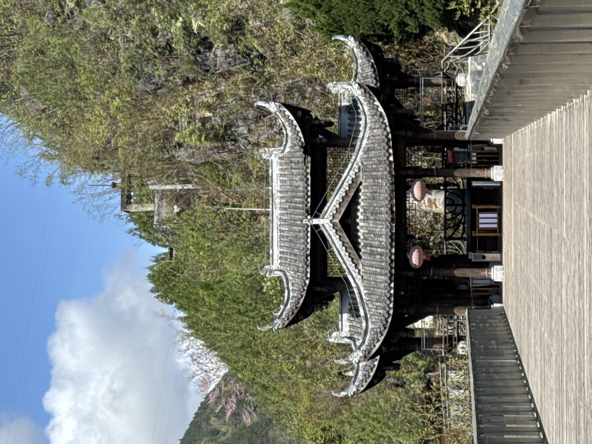 An architectural gateway with a tiled roof featuring upward-curving eaves stands against a backdrop of lush green foliage and partially visible sky in Qianjiang, China. To the right of the gateway, there's a wooden walkway or deck leading into the structure, with steps and what appears to be a handrail visible. The gateway itself is decorated with ornate, curving details and appears to lead into a building or enclosed space. The surrounding landscape is a mix of trees and vegetation, with a hint of a mountainous area in the background.