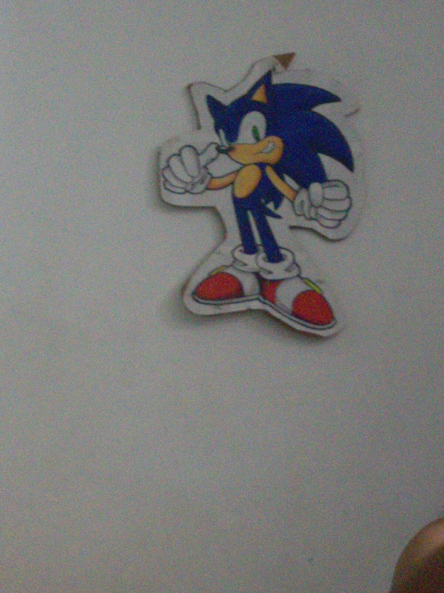 A slightly blurred image shows a cut-out of the video game character Sonic the Hedgehog affixed to a light gray wall. Sonic is depicted in his classic pose, standing with fists clenched. The background is indistinct, the image was taken at an angle, the location and origin of the image are unknown.