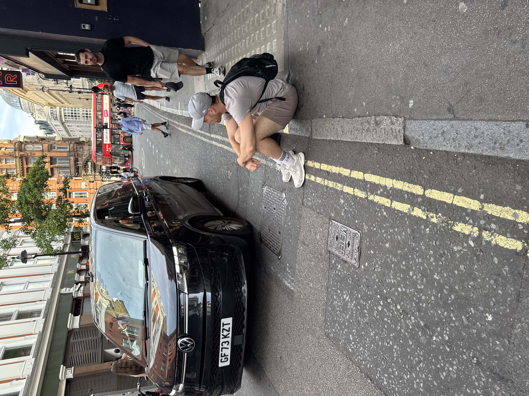 A man wearing a grey t-shirt, beige shorts, a grey baseball cap, and white sneakers is sitting on the curb of a street in London, United Kingdom, with a black backpack on his back. He is facing to his right, looking away from the camera. In front of him, there is a black Volkswagen van parked on the street. The street has yellow double lines painted on it. The background shows buildings, a red double-decker bus, and other pedestrians walking on the sidewalk. The scene appears to be a busy urban environment.