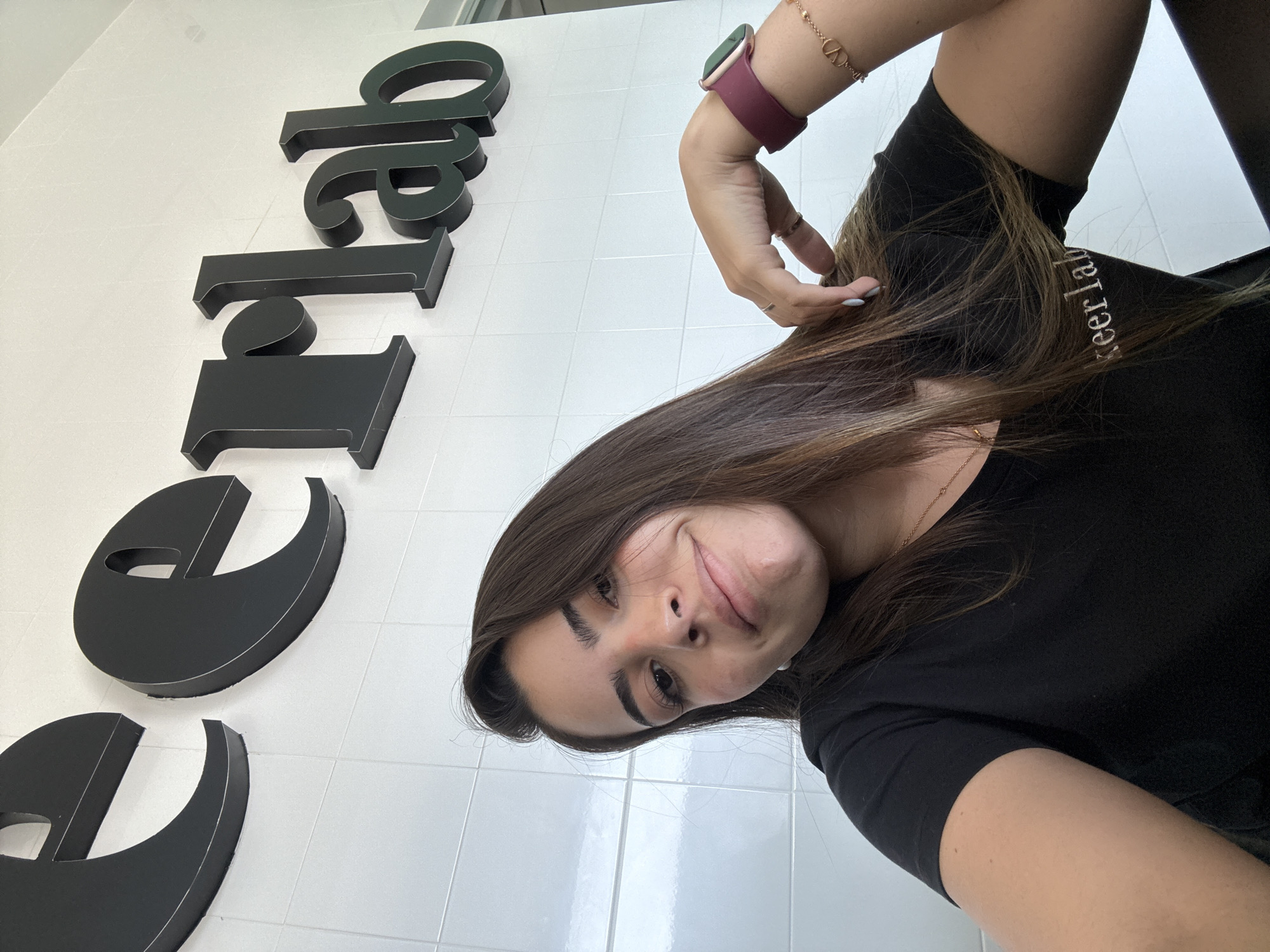 An angled, close-up selfie captures a young woman standing in front of a white tiled wall with large, dark-colored dimensional letters. The woman, with long brown hair, is looking directly at the camera with a slight, playful smirk. She is wearing a black short-sleeved t-shirt, a maroon-banded smartwatch on her left wrist, and a gold bracelet featuring a "V" or "VV" logo. Several rings adorn her fingers, and some of her fingernails are painted a light blue.
The background consists of a bright white wall covered in square tiles. Attached to this wall are prominent black 3D letters. When the image is viewed with the woman upright (rotated 90 degrees counter-clockwise), these letters spell out "BALEER" from left to right. Additionally, white text is visible on the shoulder of the woman's black t-shirt, partially reading "eer lab." The setting appears to be indoors, possibly a modern commercial or institutional space, located in San Bernardino Tlaxcalancingo, Mexico. The lighting suggests it is daytime.