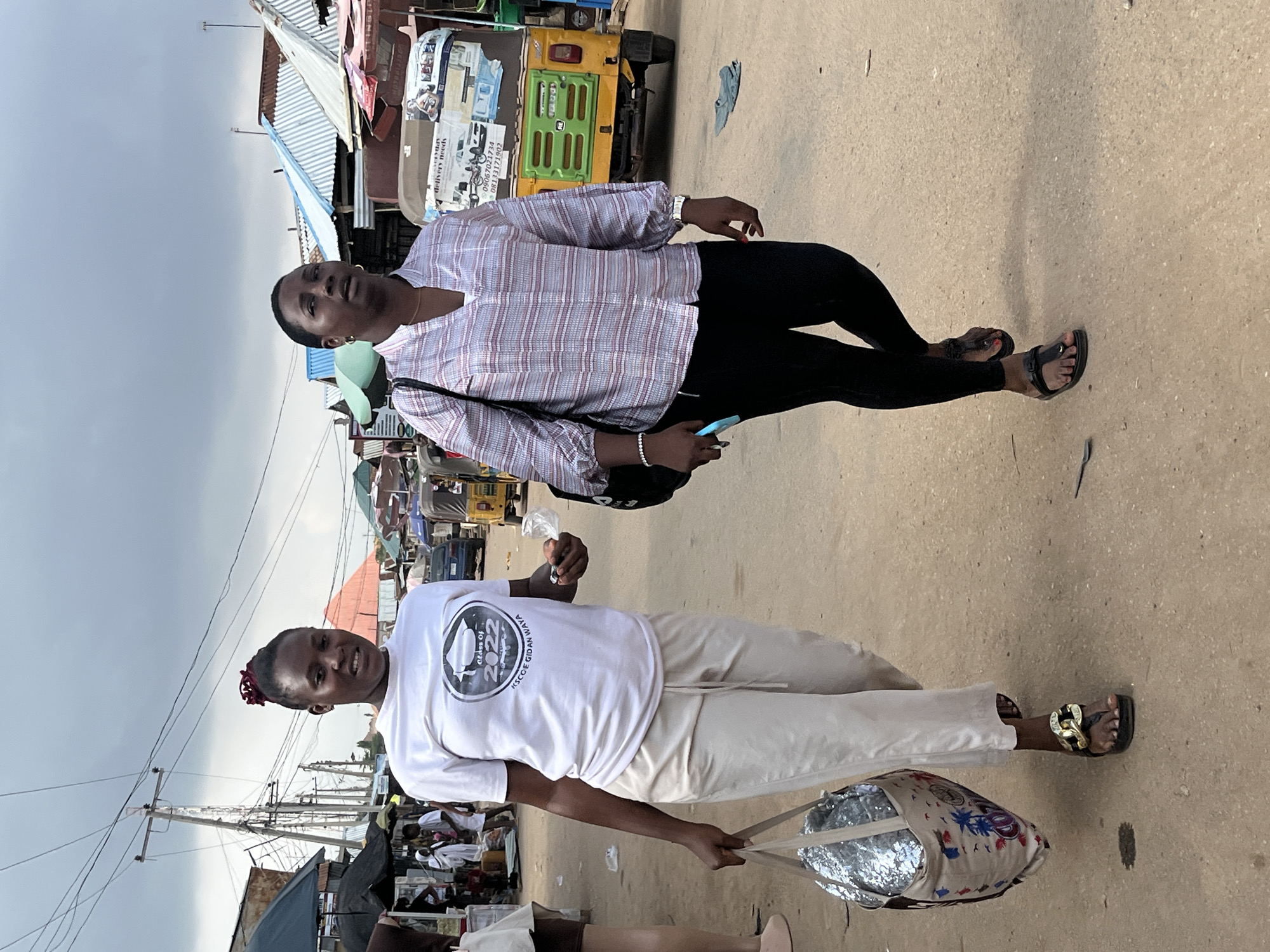 Two women are walking side-by-side on a dirt road in an outdoor market. The woman on the left is wearing a white t-shirt with a graphic that includes a graduation cap and the text "Class of 2022" and "RSCOE GIDAN WAYA", light-colored loose-fitting pants, and black platform sandals with gold accents. She is carrying a large, sequined tote bag in her left hand and a small plastic bag containing an item in her right hand. The woman on the right is wearing a striped long-sleeved blouse with a V-neck, black leggings, and black thong sandals. She is carrying a black shoulder bag and holding a light blue object, possibly a phone, in her right hand. In the background, there are market stalls with colorful awnings, power lines, a yellow and green motorized tricycle (Keke Napep), and other people. The sky is overcast. This scene is consistent with a market environment in the city of Abuja, Nigeria.