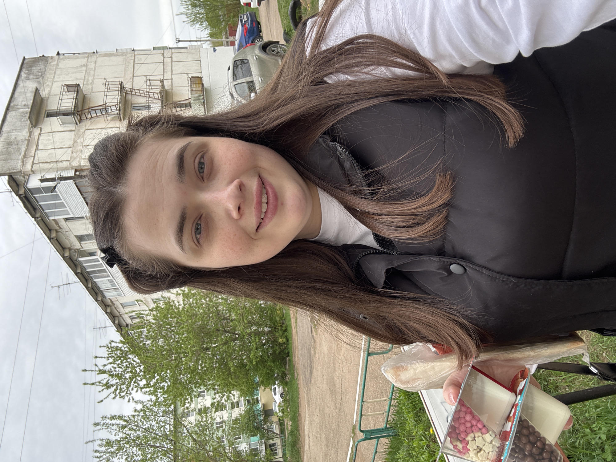 A young woman is taking a selfie outdoors, likely in a residential area. She is wearing a white shirt and a black vest. She is smiling. In her hands, she holds a box of decorative candies and a bag of something else, possibly bread.
Behind her is a building with multiple floors and fire escapes. There are trees and parked cars visible in the background. Based on the architectural style and the presence of fire escapes, the location is likely in the city of Chaikovsky, Russia.
