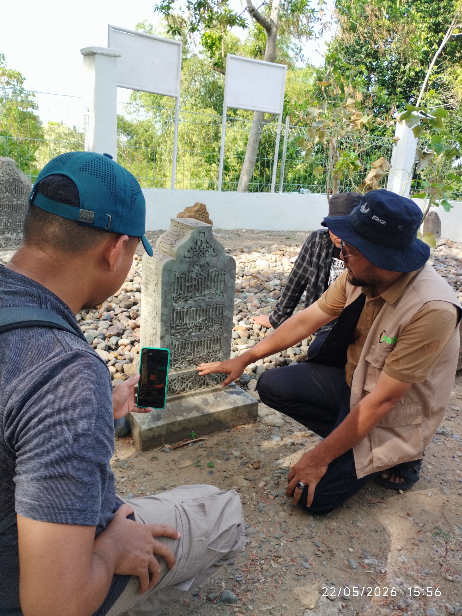 Three men are crouched around a weathered, stone headstone with Arabic script in an outdoor setting. One man, wearing a blue baseball cap and a gray shirt, is taking a photo of the headstone with his phone. Another man, wearing a tan vest and a blue bucket hat, is pointing to the headstone. A third man is crouched behind him wearing a black and white plaid shirt. The ground is covered in pebbles. The date 22/05/2026 15:56 is visible in the lower right corner of the image. The location is Ulee Tutue, Indonesia.