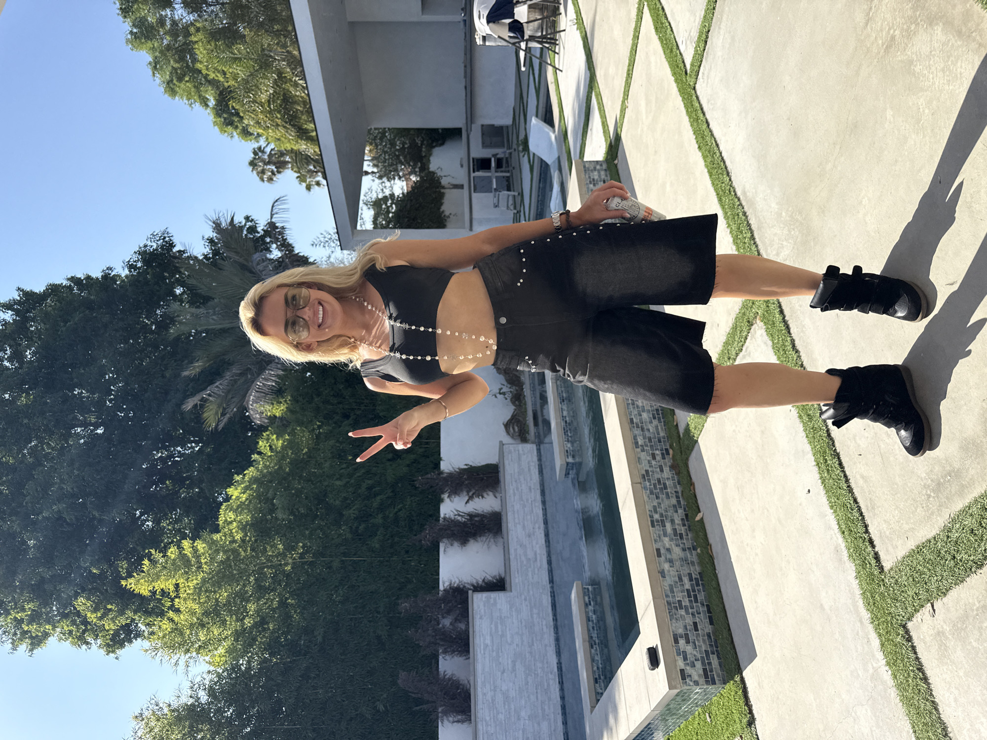A full body shot of a blond woman standing on a concrete patio with artificial turf in Los Angeles, California, USA. She is angled slightly towards the camera and is wearing a black crop top, black shorts, black boots, and sunglasses. She is smiling, facing the camera, and holding up a hand making a "peace" sign. She is also wearing a beaded necklace, a bracelet, and holding a drink in her right hand. Behind her is a swimming pool and lush greenery, suggesting a backyard setting. In the background, there is a modern house with white walls and a covered patio area. The sky is clear and blue.