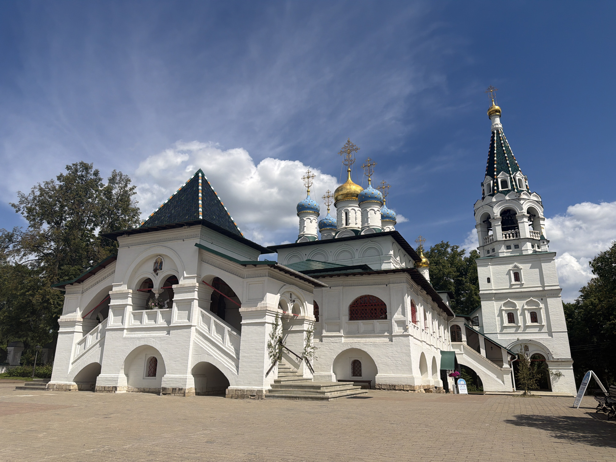 This eye-level, wide shot showcases a grand Orthodox church complex in Pavlovskaya Sloboda, Russia, under a clear blue sky dotted with white, fluffy clouds. The primary subject is a multi-part white architectural structure, characteristic of traditional Russian design, bathed in bright daylight that casts distinct shadows on the paved courtyard.
The complex features a prominent section on the left with white walls, arched openings, and large staircases leading to an elevated platform, all topped with a dark green roof patterned with colorful tiles. The main church building, central in the image, boasts multiple vibrant blue onion domes alongside a larger golden dome, each crowned with ornate golden Orthodox crosses. To the right, a tall, slender bell tower rises, matching the white facade of the church, with a distinct green and white patterned spire also topped with a golden cross. The tower includes several arched openings and smaller windows.
The ground is paved with light-colored rectangular blocks, suggesting a spacious plaza or courtyard. Lush green trees frame the complex on both sides, providing a natural backdrop. There are no people visible, suggesting a tranquil moment. On the paved area near the bell tower, two white A-frame signs are present; one partially visible sign appears to read "Свято-Духовский...", likely indicating part of the complex's name, while the other is less legible. The overall impression is one of architectural beauty and historical significance in serene surroundings.