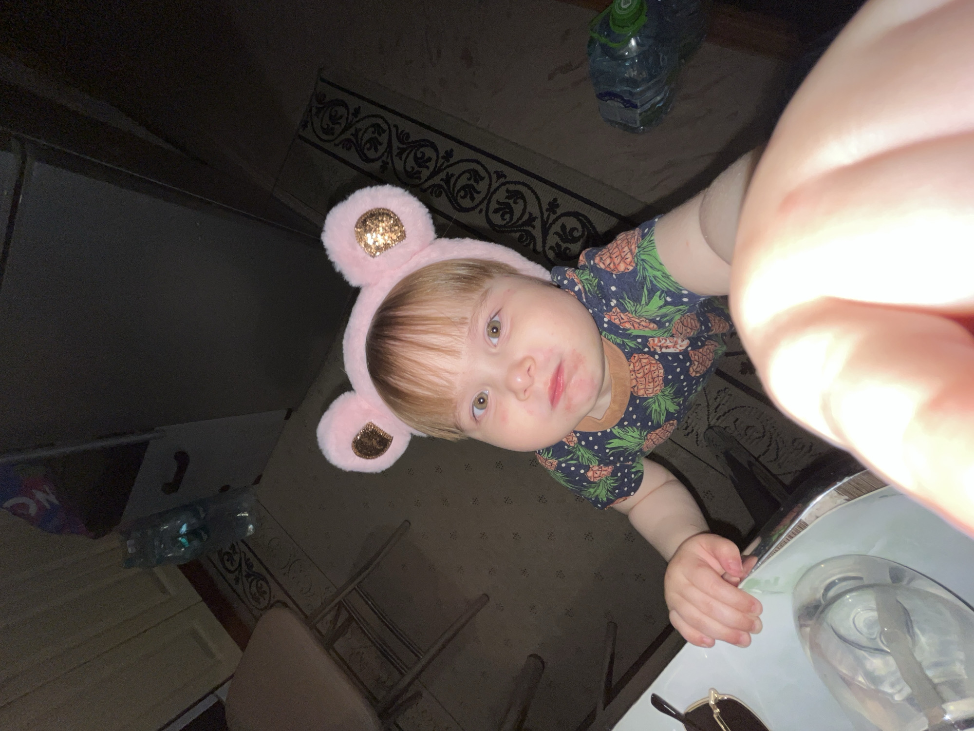 A child with light brown hair is wearing a pink headband with sparkly bear ears. The child is wearing a blue shirt with a pineapple print. The child is standing near a table with a clear dispenser, possibly of soap or lotion, on it. A pair of sunglasses is on the table near the dispenser. A bottle of water is visible in the background, which also includes a dark ornate rug and wooden chairs. The location is Unknown, Unknown.