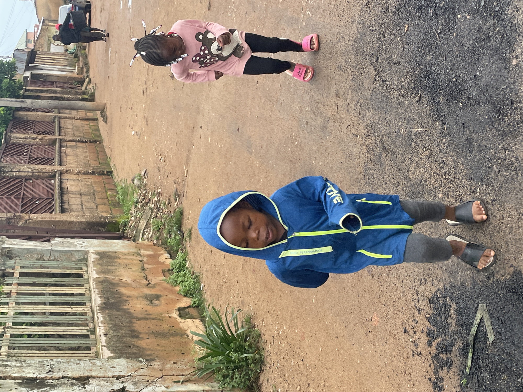 A boy wearing a blue hooded jacket and grey leggings walks on a dirt road in Jos, Nigeria. He is looking down and to the left. A girl in a pink long-sleeved shirt with a bear graphic and black leggings walks in the background, also looking down. A fence made of vertical bars is on the left side of the road, with a rough, weathered wall beneath it. Buildings are visible in the background.