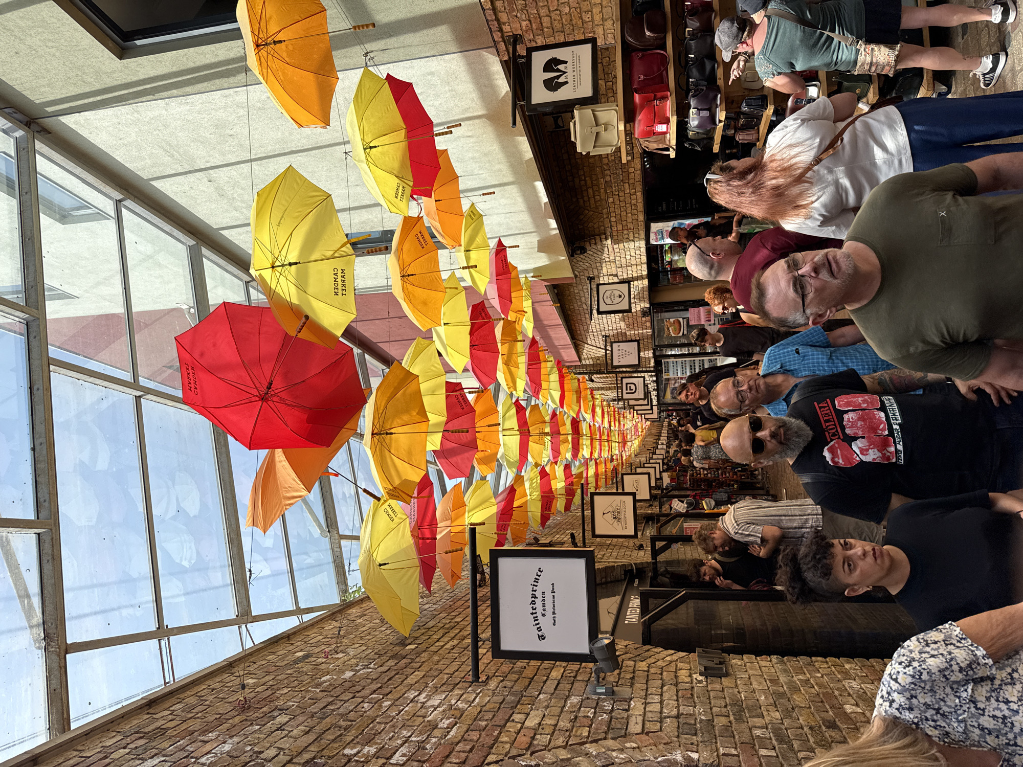 This photograph depicts a street scene with various individuals and an overhead display of colorful umbrellas. The location appears to be a shopping district, given the store displays and signage.
Overhead, a multitude of umbrellas in red, orange, and yellow are suspended, creating a visually striking canopy over the pedestrian walkway. These umbrellas are evenly spaced and create a tunnel-like effect, leading the viewer's eye down the street.
Along the right side of the image, pedestrians are strolling and browsing in front of shops. Several people are captured in various poses, suggesting they are either shopping or simply passing through the area. Store displays are visible behind the pedestrians, showcasing various merchandise such as handbags and other items. Signs with text are affixed to the building facades.
The buildings on both sides are constructed from red brick, which adds a rustic texture to the setting. The left side features glass panels above the brick, indicating a partial enclosure of the street.
The scene is well-lit, suggesting it is daytime, and the overall impression is a busy, vibrant urban setting. The specific location is likely a commercial area in the city of Camden, as suggested by a shop sign.