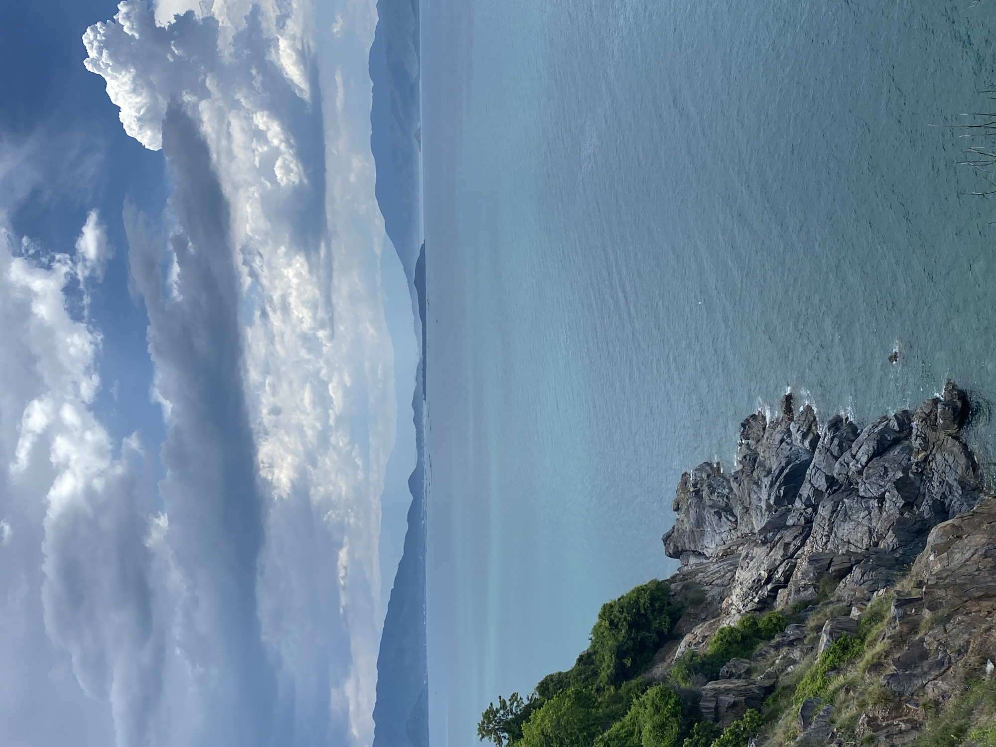 The image is a high-angle, full shot of a coastal landscape under a cloudy sky. In the foreground, a rugged, rocky cliff covered in green vegetation juts out into the sea. The cliff face shows layered rock formations and sparse green foliage. The sea is a deep turquoise blue, with gentle ripples. In the distance, a hazy outline of mountains stretches across the horizon. The sky is filled with dramatic, dark gray clouds interspersed with patches of bright white clouds and rays of sunlight breaking through. This scene is likely from a high vantage point overlooking the coast near Nha Trang, Vietnam, possibly from Hon Ba Island or a similar elevated coastal area known for its dramatic cliffs and ocean views. There are no people or any explicit actions depicted in the image.