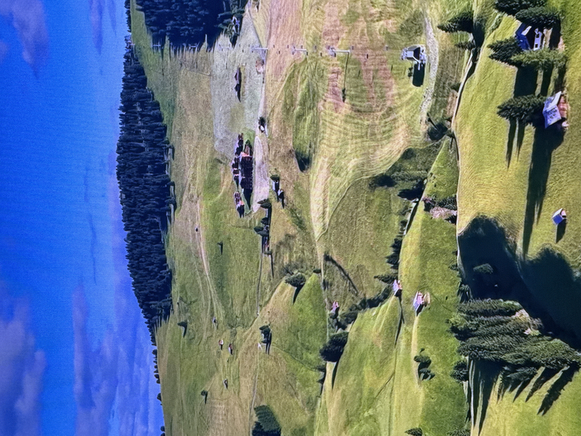 The image depicts a mountainous landscape with rolling green hills and scattered trees. Several small buildings, resembling chalets or houses, are visible nestled on the hillsides. A ski lift with several towers and cables traverses the scene, suggesting a ski resort or a mountain recreational area. The sky is a vibrant blue with some scattered clouds. There are no visible people or any elements that would indicate the location is Dubai, United Arab Emirates. The terrain is consistent with alpine or mountainous regions.