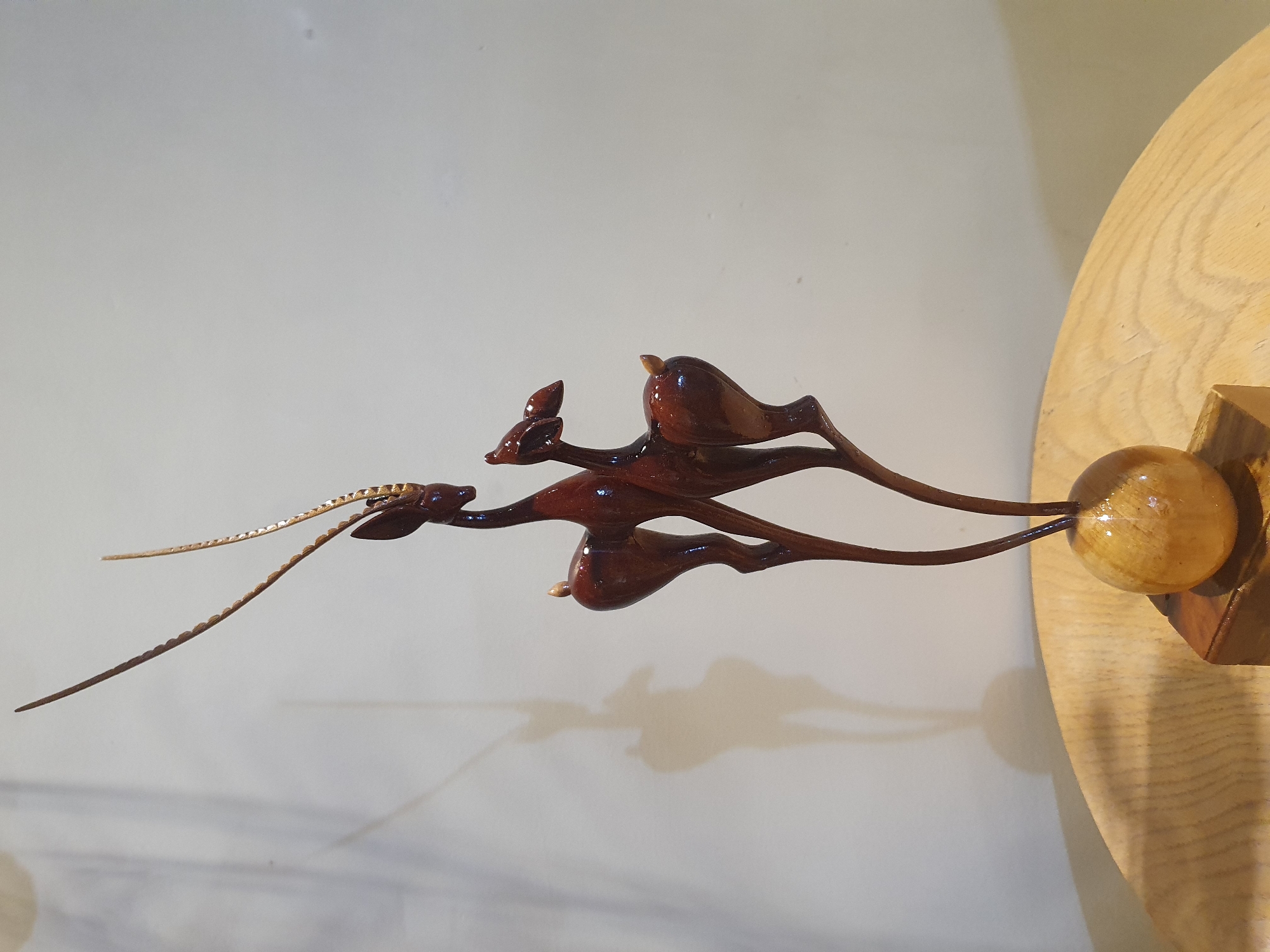 A polished, dark brown wooden sculpture of two gazelles is displayed on a light-colored wooden table. The sculpture depicts one larger gazelle with long, spiraling horns and a smaller gazelle standing beside it. The gazelles are carved with slender bodies and delicate legs, conveying a sense of grace and movement. The sculpture is mounted on a spherical wooden base, which is itself placed on a rectangular wooden block. The background is a plain, off-white wall, with a soft shadow of the gazelles cast upon it. The overall lighting is soft, highlighting the smooth texture and rich color of the wood. There is no indication of the location in Shiraz, Iran, or any people or actions.
