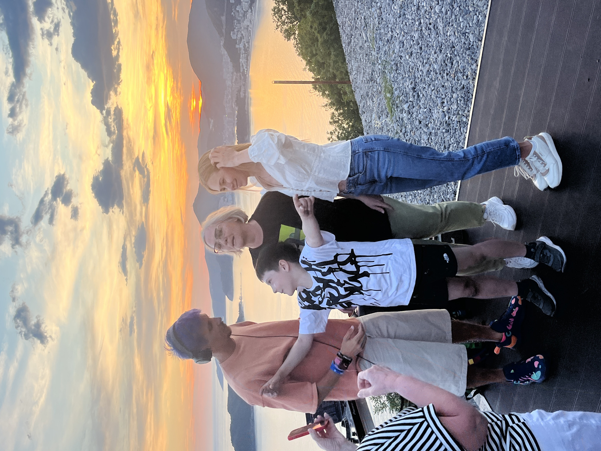 A photo taken outdoors, rotated 90 degrees, shows a group of five people standing on a dark brown deck with a view of what appears to be a sunset over water. The background reveals a city landscape along the coast, possibly the city of Budva in Montenegro, given the mountainous terrain and coastal setting.
The uppermost person in the photo is a woman with long blonde hair, wearing a white blouse and blue jeans, with white sneakers. Below her is an older woman with short grey hair, wearing glasses and a black top. Next is a young boy in a white t-shirt with a black pattern and black shorts, with dark sneakers. The next person down is wearing an orange shirt, beige shorts and socks with a colorful pattern, with a purple-colored hair. At the bottom of the image, a person wearing a white and black striped shirt is partially visible.
The sky is filled with orange and yellow hues suggesting either sunrise or sunset, with clouds scattered across the sky. A body of water is visible in the background, with land or mountains on the opposite side. The deck they are standing on is made of dark brown planks. To the right of the deck, a gravel-covered area with some greenery is present.