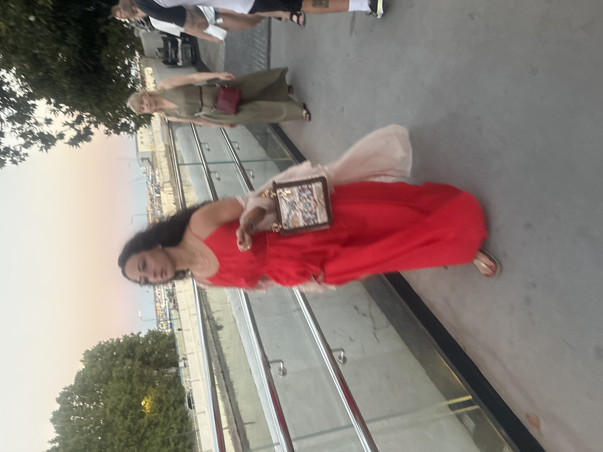 This media file, oriented vertically, captures a street scene with several individuals. In the foreground, a woman in a vibrant red dress is walking towards the viewer, holding a decorative clutch bag. Her dark hair is flowing, and she appears to be posing for the photo. Behind her, another woman in a green dress is walking away, also carrying a red bag. Further in the background, partially obscured, are other people. The scene appears to be an outdoor walkway or promenade with metal railings and a paved surface. Trees and some buildings are visible in the distance, along with a body of water. The sky suggests it might be late afternoon or early evening, with a soft, diffused light. The overall atmosphere seems casual and relaxed, possibly a tourist or leisure setting in Sochi, Russia. There is no visible text in the image.