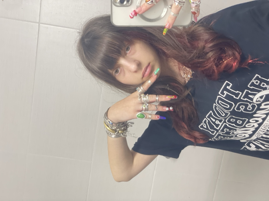 A young woman with brown hair and bangs poses in a mirror selfie, holding her phone in her right hand. Her nails are long and decorated with various colorful designs. She wears numerous rings on both hands and a stack of bracelets on her left wrist. She's wearing a black t-shirt with white text. The background appears to be a tiled wall, possibly in a bathroom or public space.