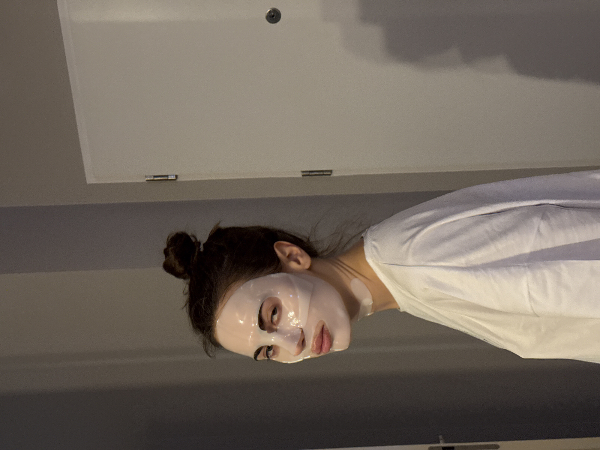 A woman with her hair in a bun is wearing a white sheet mask and a white t-shirt. She is in an indoor environment, likely a bathroom, as indicated by the flat, white ceiling panel with a metal knob and hinge in the background. The walls are painted in a neutral, light gray tone. The location is Unknown, Unknown.