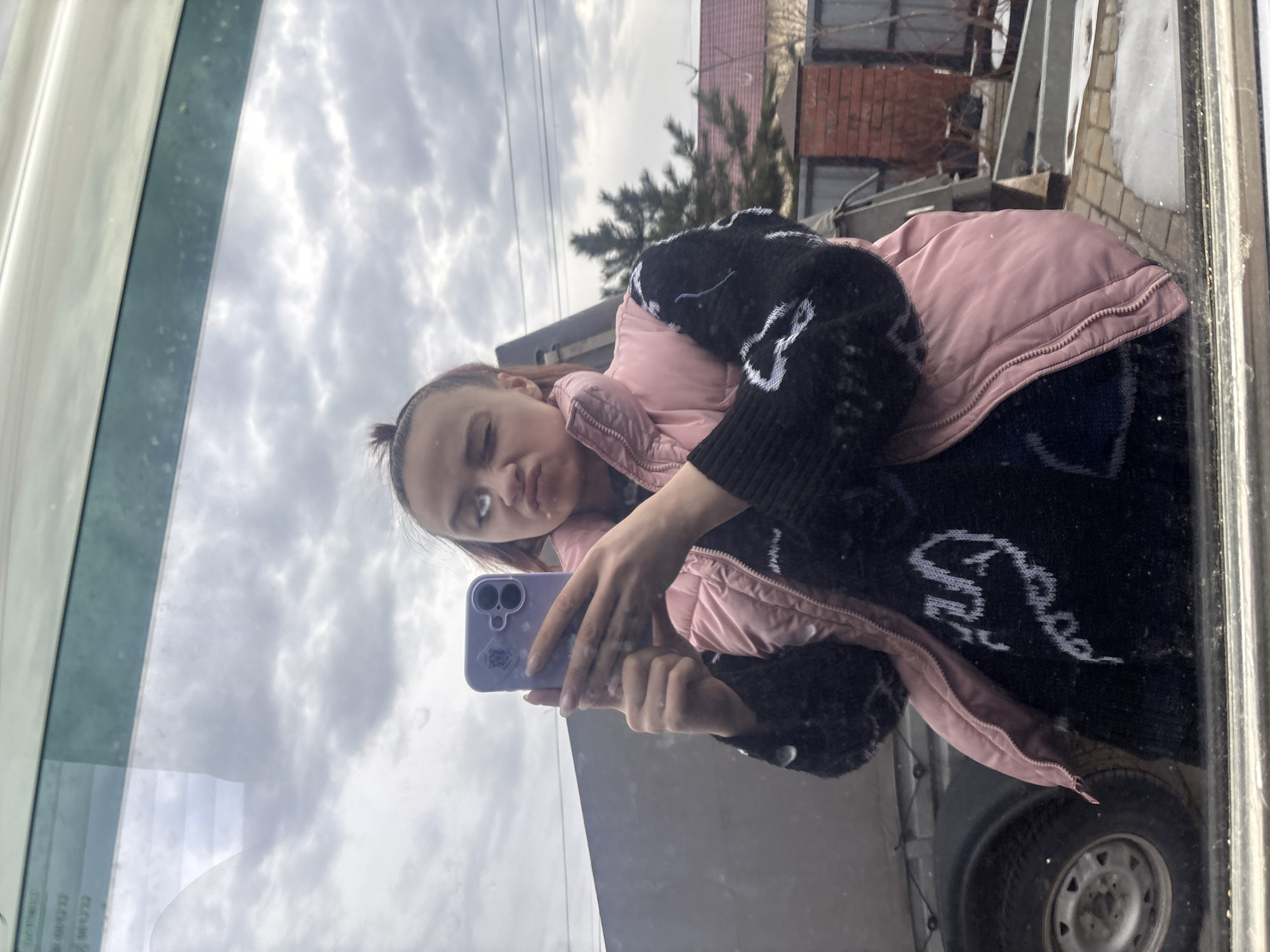A young woman is taking a selfie in Прикамский, Russia. She is wearing a pink vest over a black sweater and holding a purple smartphone. The reflection in the glass shows the sky, clouds, a building with a red roof, a tree, and part of a vehicle, including a tire.
