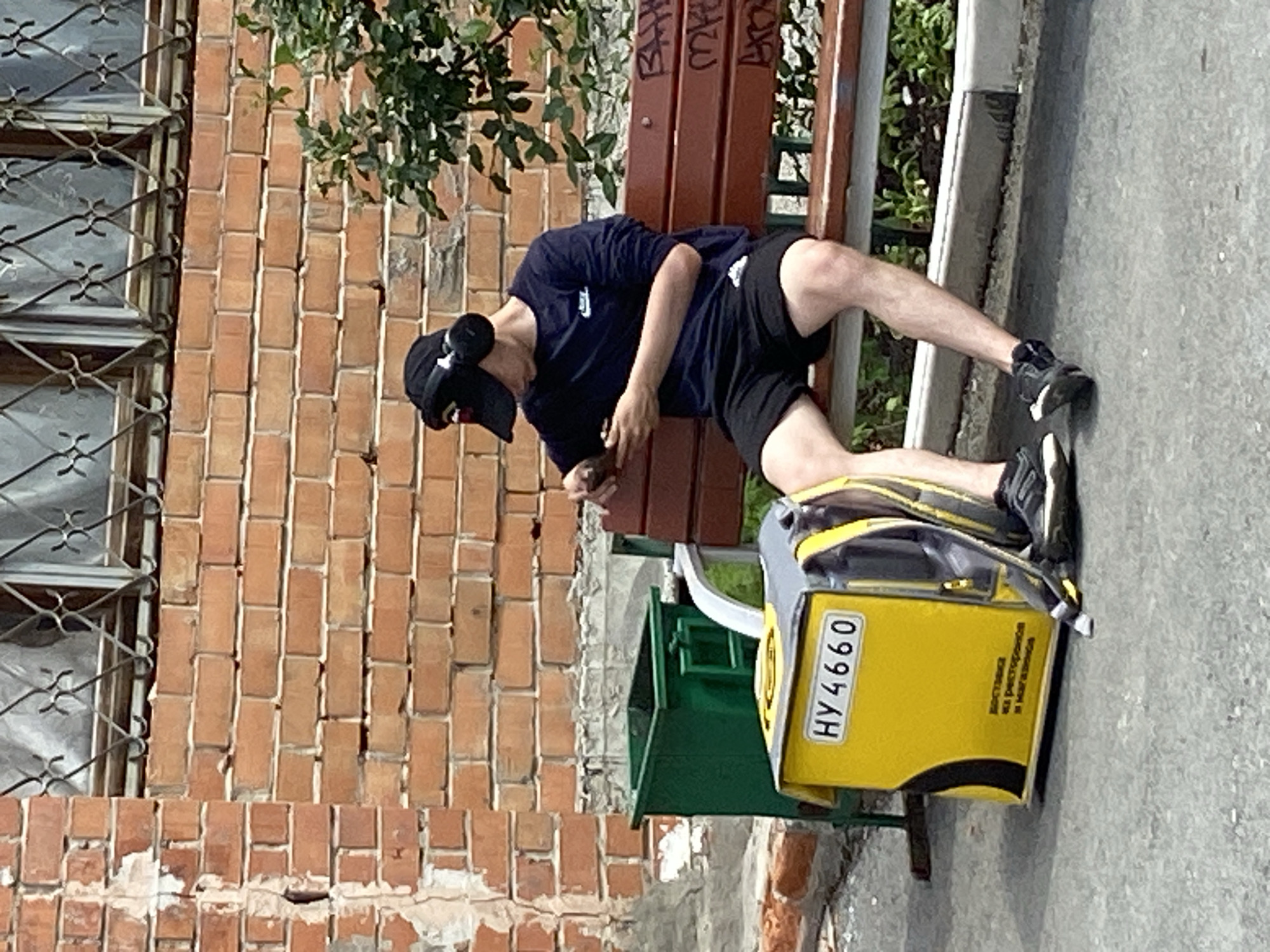 A young person wearing a black baseball cap and headphones sits on a wooden bench, looking at a smartphone. They are wearing a dark blue long-sleeved shirt and black shorts. Beside them on the ground is a bright yellow delivery backpack with "HY4660" printed on a white rectangular plate, and some Russian text. A green trash can is to the left of the backpack. The background consists of a brick wall with barred windows and some greenery. The scene appears to be an outdoor urban setting.