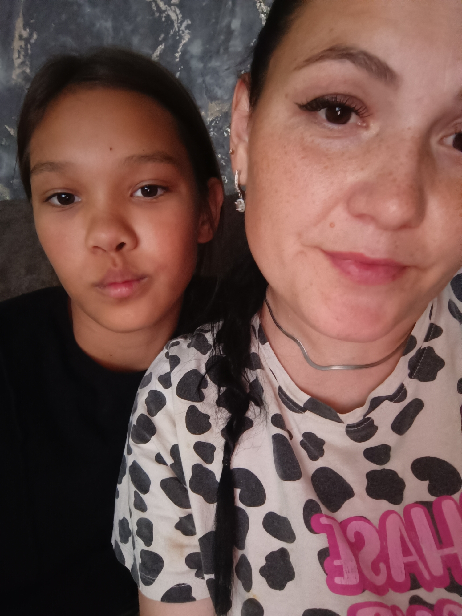 The image is a close-up selfie of two females. The female on the left appears to be a younger girl with brown hair, brown eyes, and is wearing a black t-shirt. The female on the right is an adult with dark hair, brown eyes, fair skin with freckles, and is wearing a cow-print shirt and a thin silver necklace. The background is a dark, textured wall. The image does not provide any information or visual cues to indicate its location in the city of Karsha, Russia.