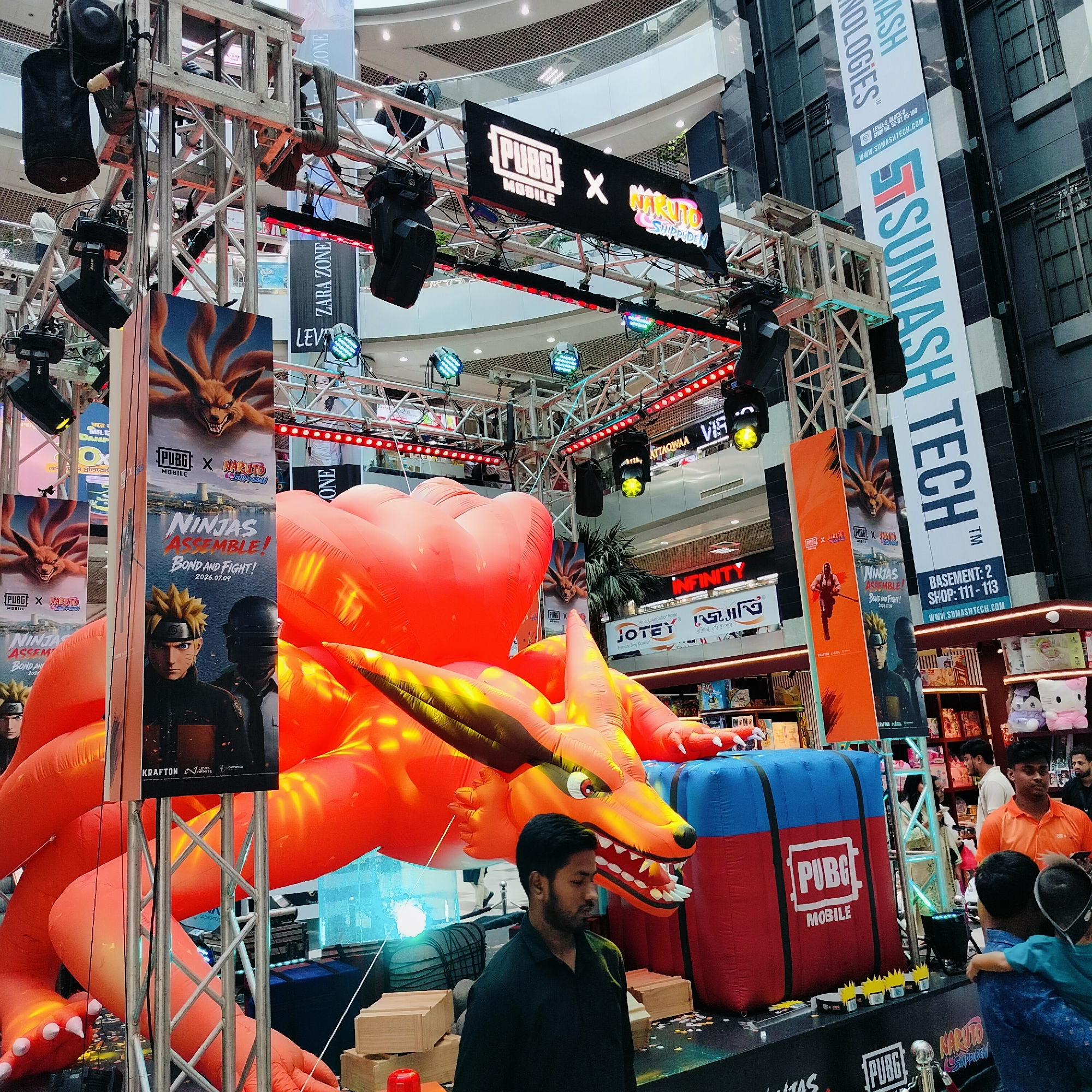 The image captures an indoor event setup, likely in a shopping mall, featuring a large inflatable installation of a fox-like creature, possibly Kurama from Naruto, in a dynamic, roaring pose. The creature is predominantly orange with yellow accents, and its mouth is agape, revealing sharp teeth. A large blue and red inflatable cube with the "PUBG MOBILE" logo rests beside it.
Above the inflatable, a black banner displays "PUBG X NARUTO SHIPPUDEN" in illuminated white text and logos. This banner is suspended from a metal truss system equipped with various stage lights, some emitting red and green hues.
Several vertical banners are visible, one prominently featuring the "PUBG X NARUTO SHIPPUDEN" theme with images of characters and text like "NINJAS ASSEMBLE! BOND AND FIGHT!". Another vertical banner to the right displays "SUMASH TECH" in large blue letters, with smaller text indicating "LEVEL: 2, BLOCK: 6, SHOP NO: 02-02-05-100" and a website URL "WWW.SUMASHTECH.COM". This banner's placement suggests Sumash Tech is a tenant or sponsor at this location.
In the foreground, a man in a dark shirt stands looking down, seemingly observing the setup. Behind him, the crowd and shop displays are visible, indicating a commercial setting. In the background, the architecture of a multi-level indoor space is apparent, with railings and escalators suggesting a shopping mall environment.
The presence of "SUMASH TECH" and the typical setup of such promotional events strongly suggest this location is in Dhaka, Bangladesh, as Sumash Tech is a well-known electronics retail chain in the country. The overall scene depicts a promotional event for the game "PUBG MOBILE" in collaboration with "Naruto Shippuden", utilizing eye-catching inflatables and banners within a public indoor space.