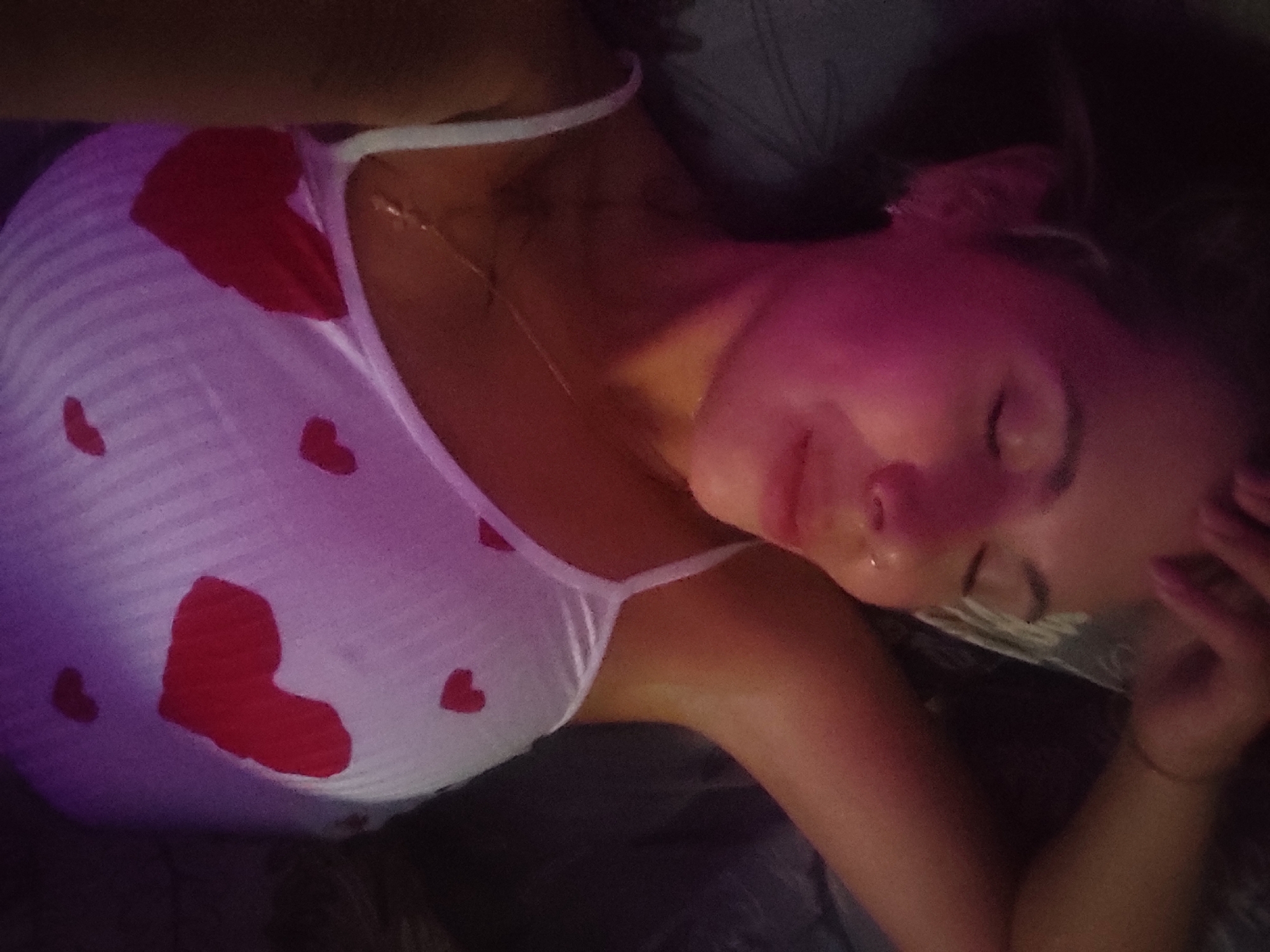 A person is lying down on a bed with a pillow under their head. The person is wearing a white tank top with red heart patterns. They have a gold chain necklace on. Their face is lit with a pinkish-purple light. The location is Chaikovsky, Russia.