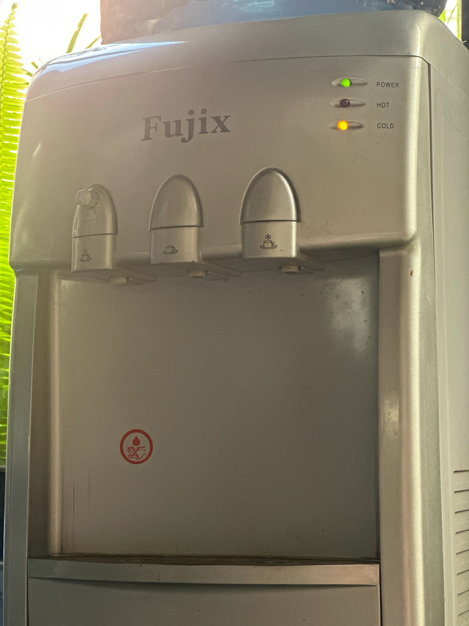 A close-up, eye-level shot captures a silver Fujix water dispenser against a softly lit, outdoor background. The dispenser has three spigots, two of which are decorated with small icons: one with a steaming cup of coffee, and another with a cup of coffee. The third spigot is on the far left and has a lever on its side labeled "ON OFF". Above the spigots, three indicator lights are visible: a green light labeled "POWER", a dark red light labeled "HOT", and a yellow light labeled "COLD". A red circular warning sticker with a droplet of water and two hands is positioned on the lower left side of the dispenser. The background features blurred green foliage, suggesting it is situated outdoors, possibly in a garden or near a courtyard in Lalitpur, Nepal.