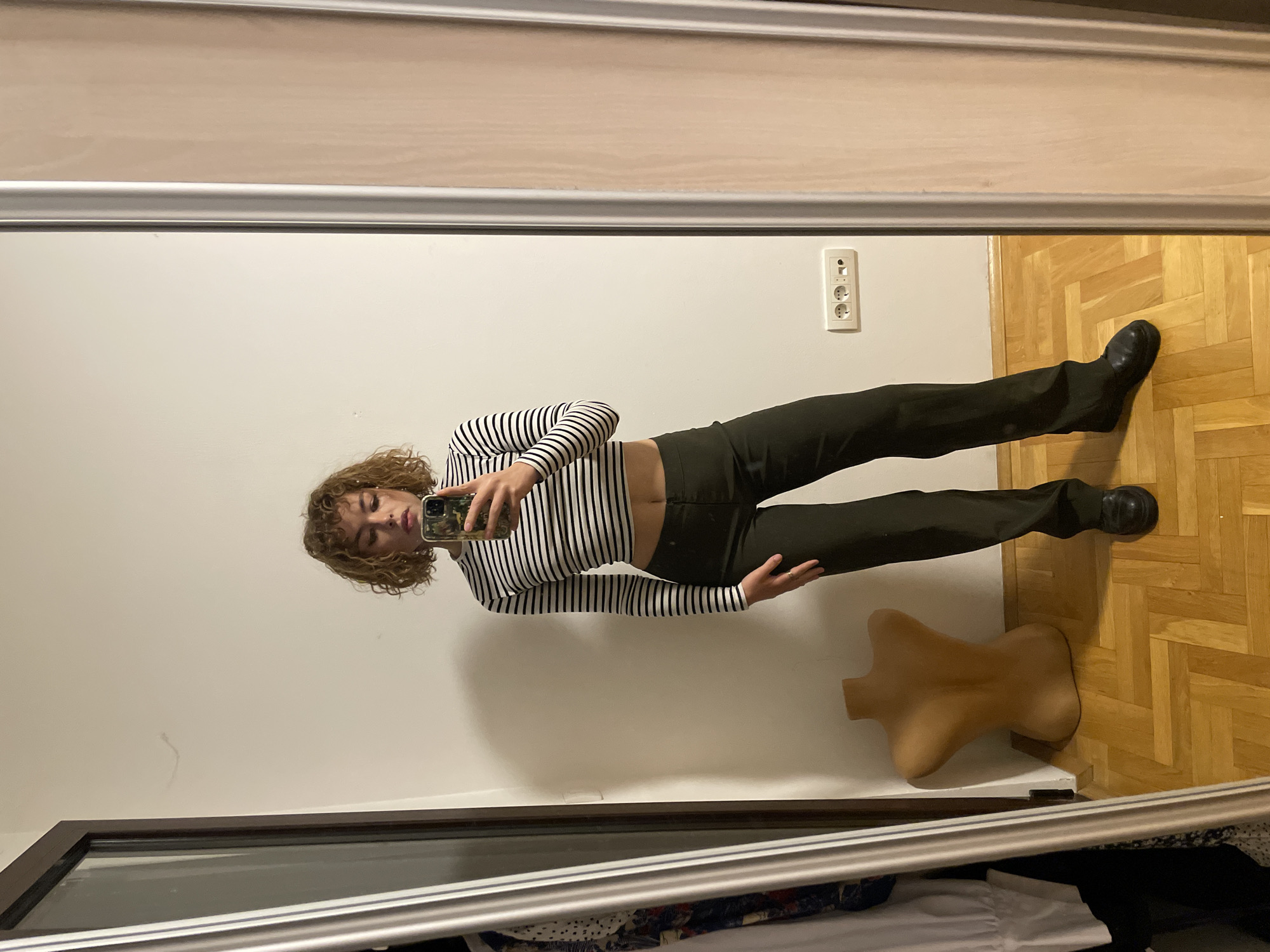 The image shows a person taking a mirror selfie in an indoor setting. The person has curly hair, is wearing a striped long-sleeve shirt, dark pants, and black shoes. They are holding a smartphone. A partial mannequin torso is visible on the floor near the person. The floor is wooden parquet. The walls are white. There is an electrical outlet on the wall. Part of a closet with clothing inside is visible at the bottom of the frame. The location (Boreti, Montenegro) cannot be determined from the image.